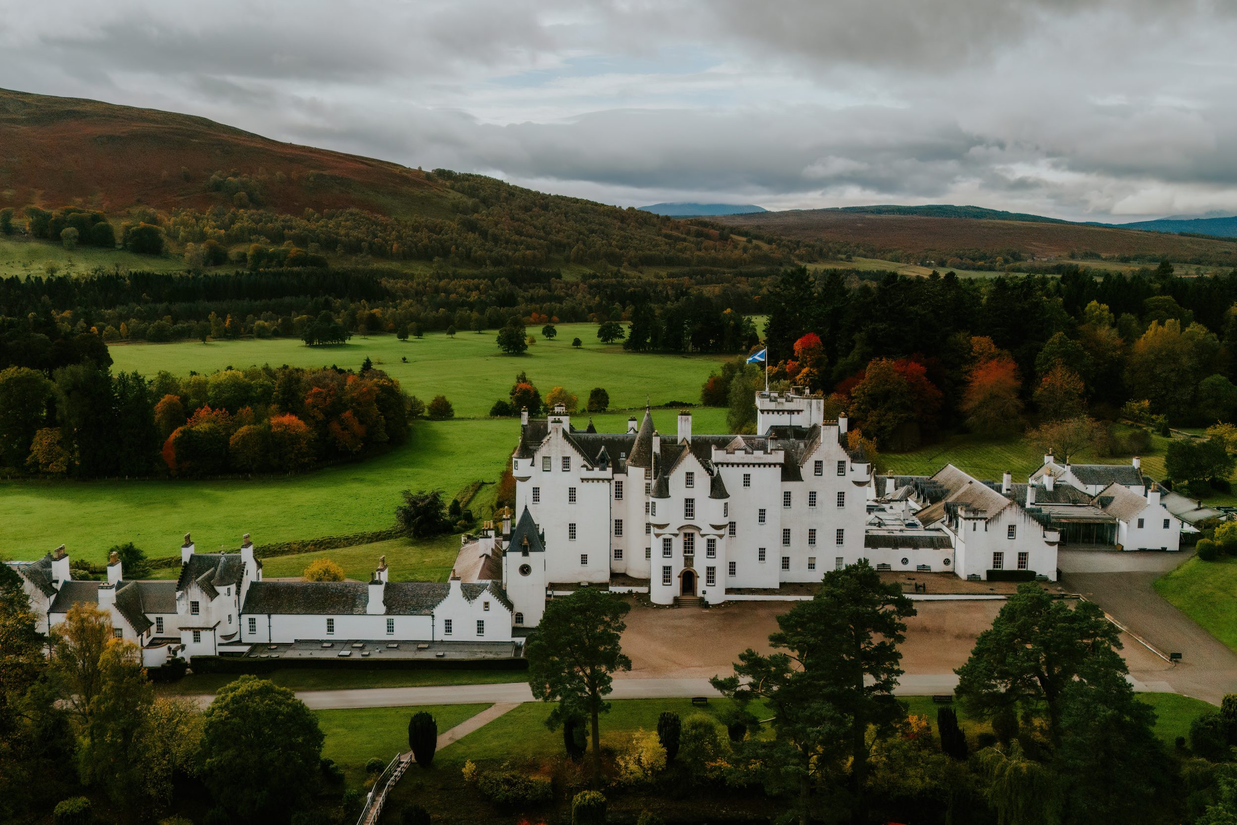 Aerial view of Blair Castle at Atholl Estate with Scottish flag, lawns and autumn hills under a cloudy sky.
