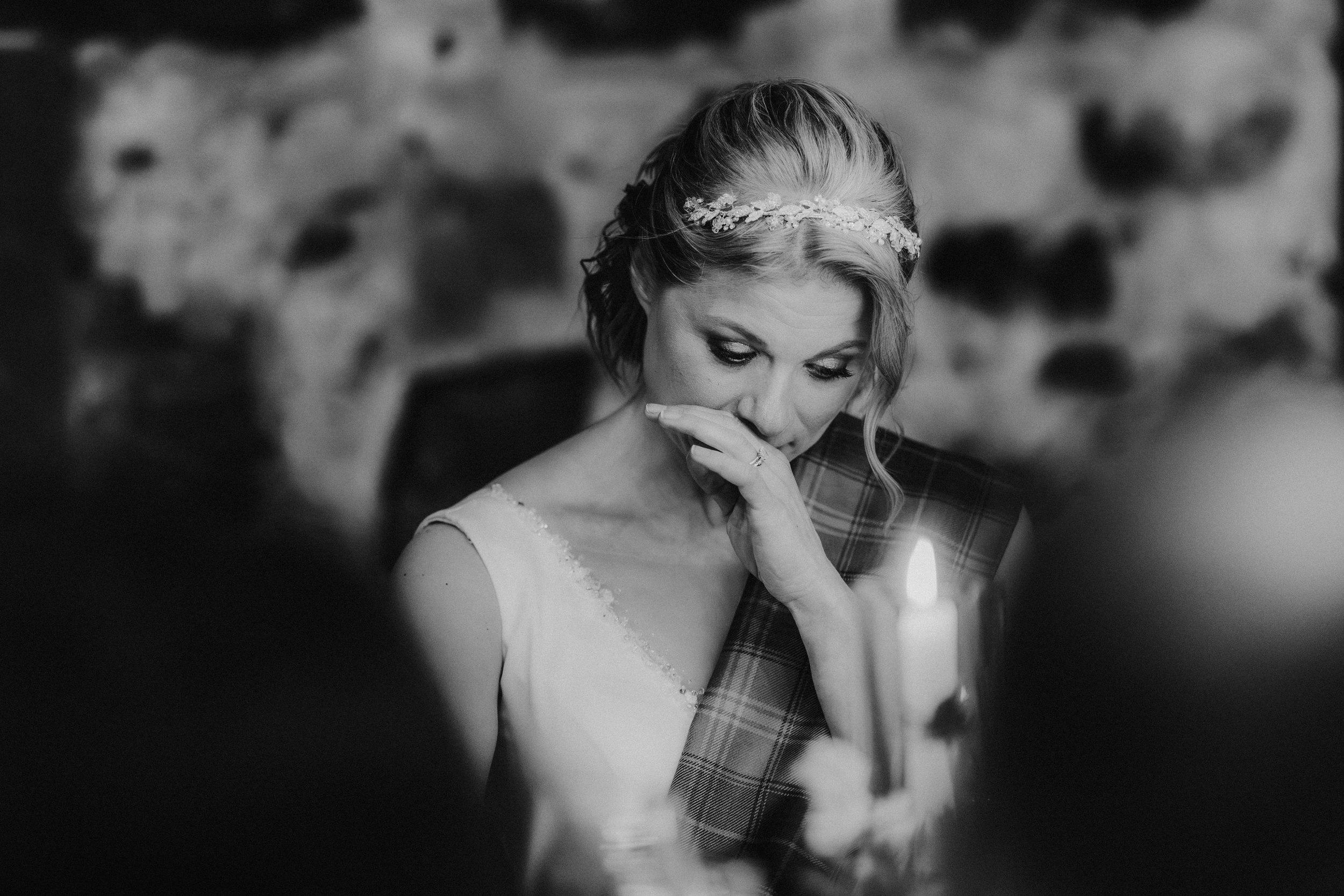 Black-and-white close-up of the bride Lizanne, hand to her face, wearing a jeweled headband and tartan sash by a candle.