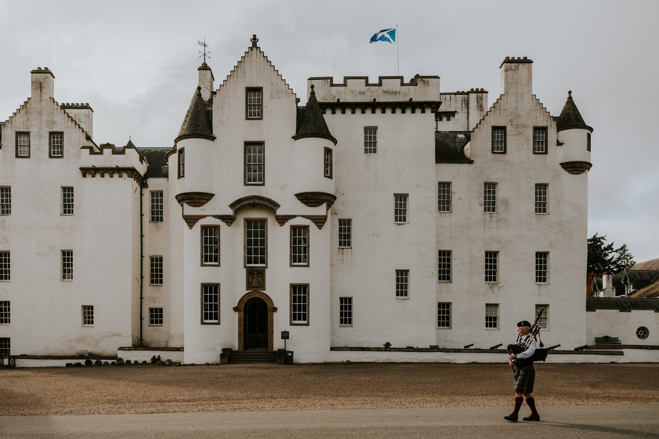 Blair Castle frontage with Scottish flag and a bagpiper in tartan walking across the courtyard.