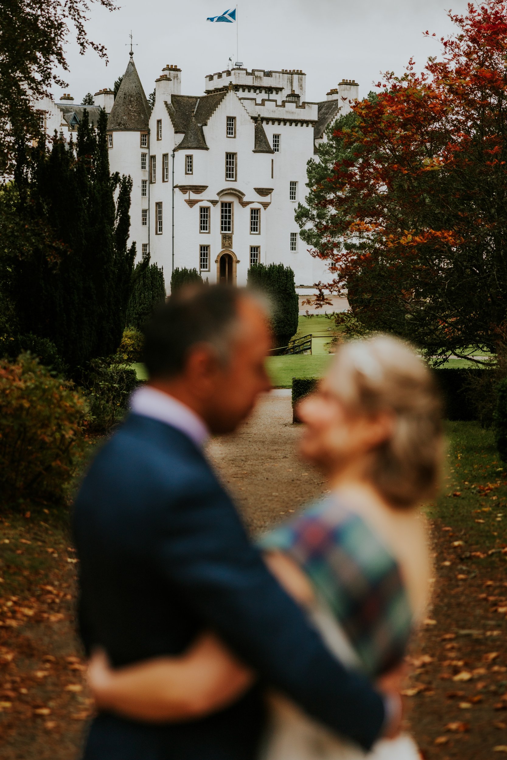 Blurred couple embracing on a path with Blair Castle and a Scottish flag framed by autumn foliage.