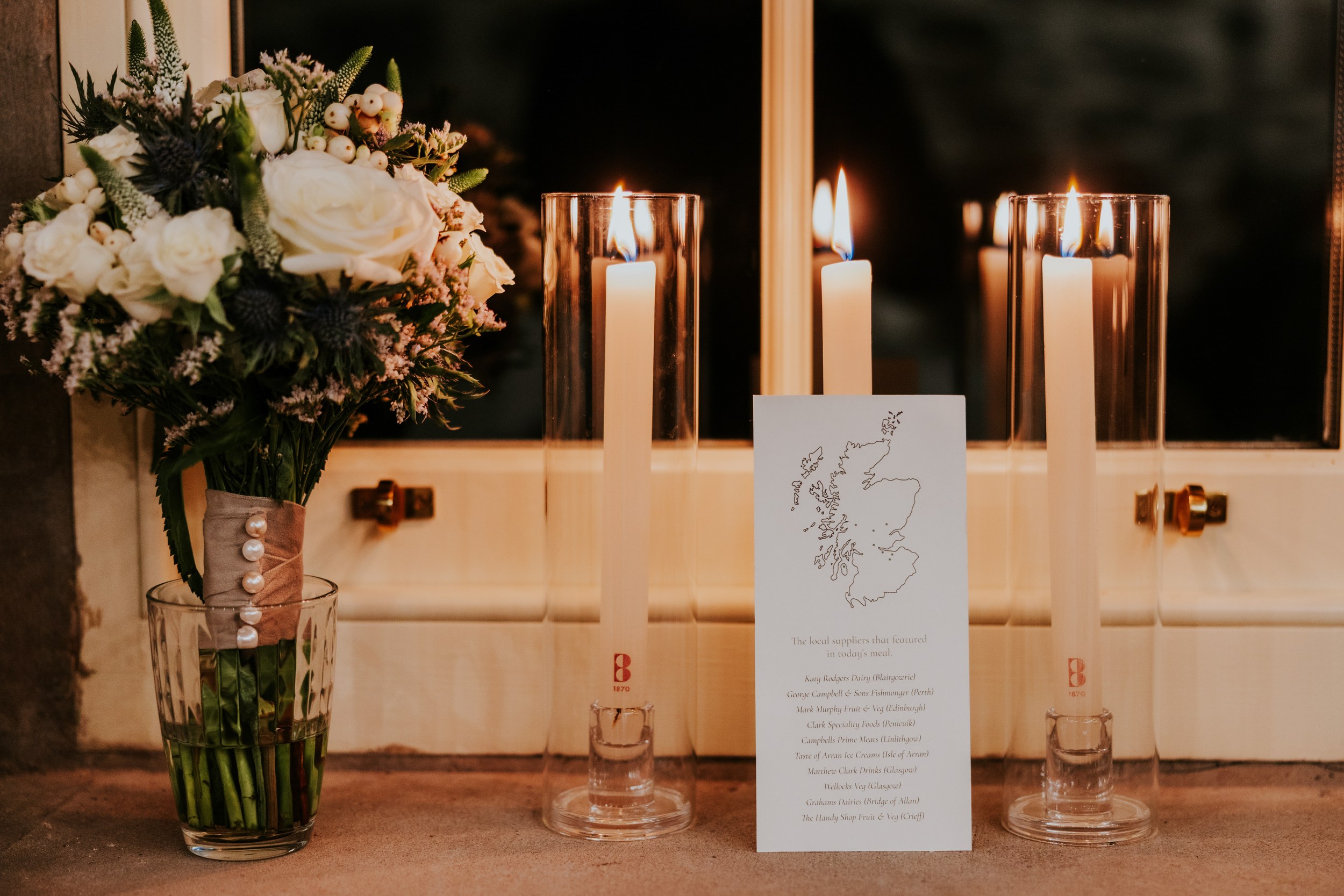 Bridal bouquet beside three lit pillar candles and a printed menu card with a map of Scotland on a windowsill.