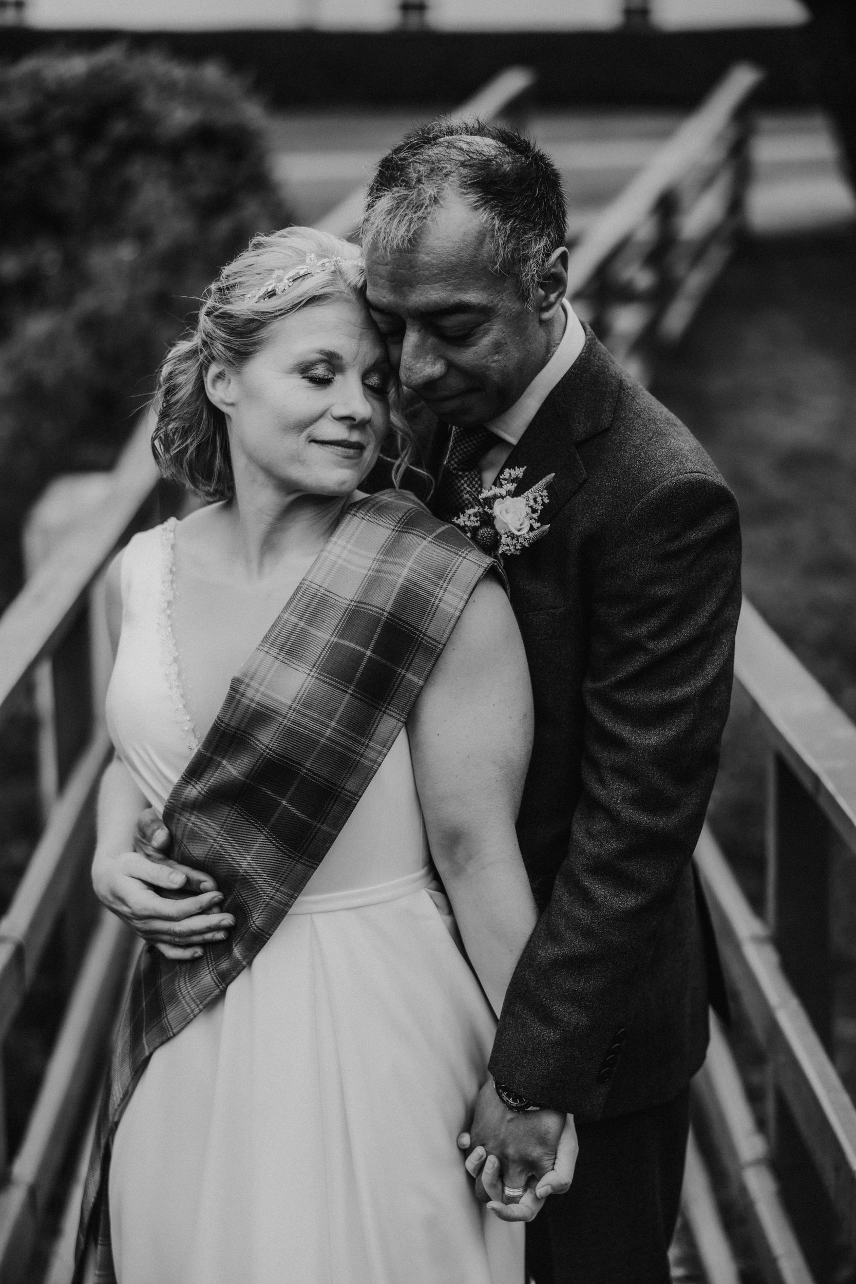 Bride and groom embrace on a wooden bridge; bride wears a tartan sash and headpiece, the groom holds her and their hands are clasped.