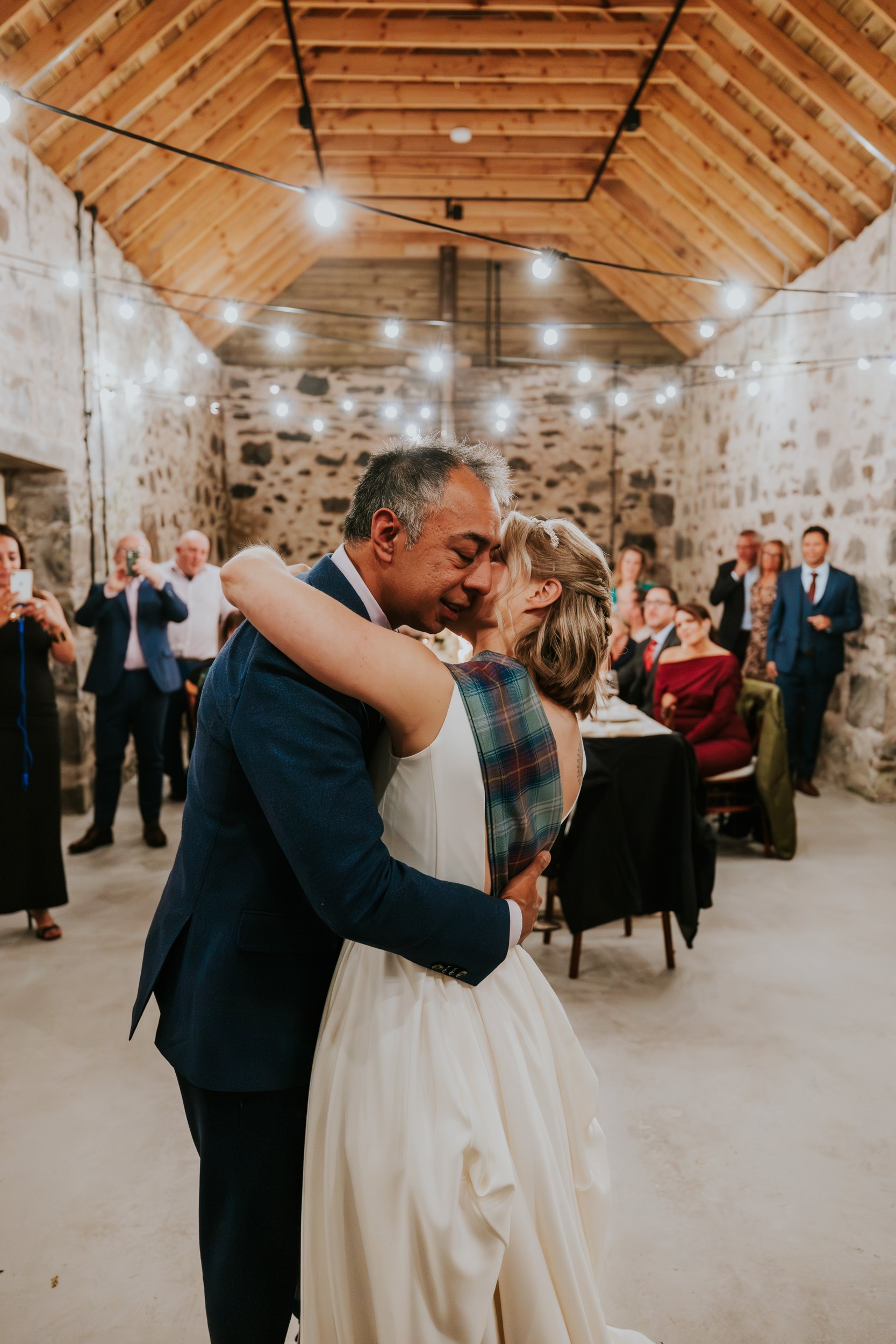 Bride and groom embrace while dancing in a stone-walled barn lit by string lights with guests watching.