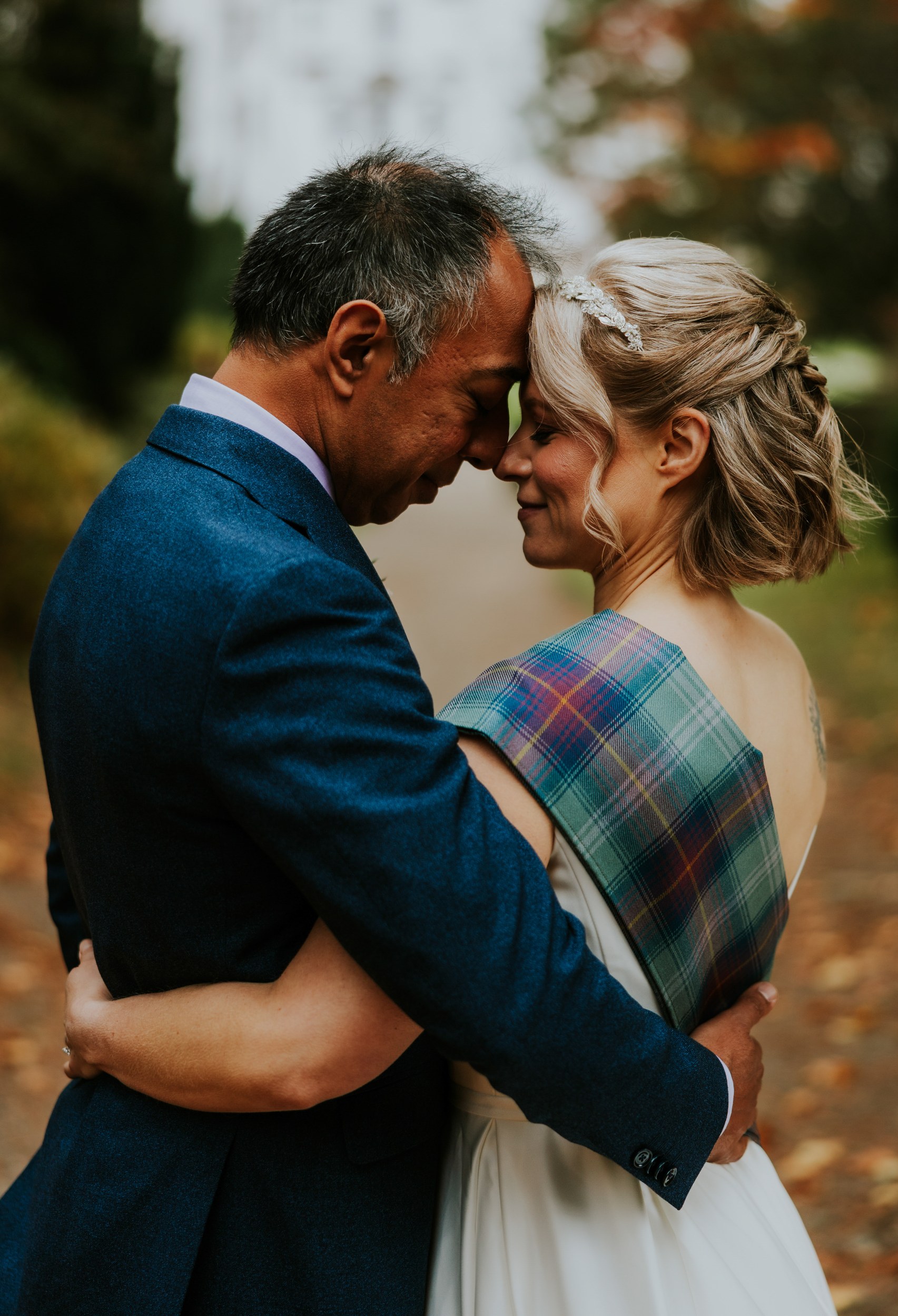 Bride and groom forehead-to-forehead embrace; groom in blue suit, bride with tartan sash and hairpiece.