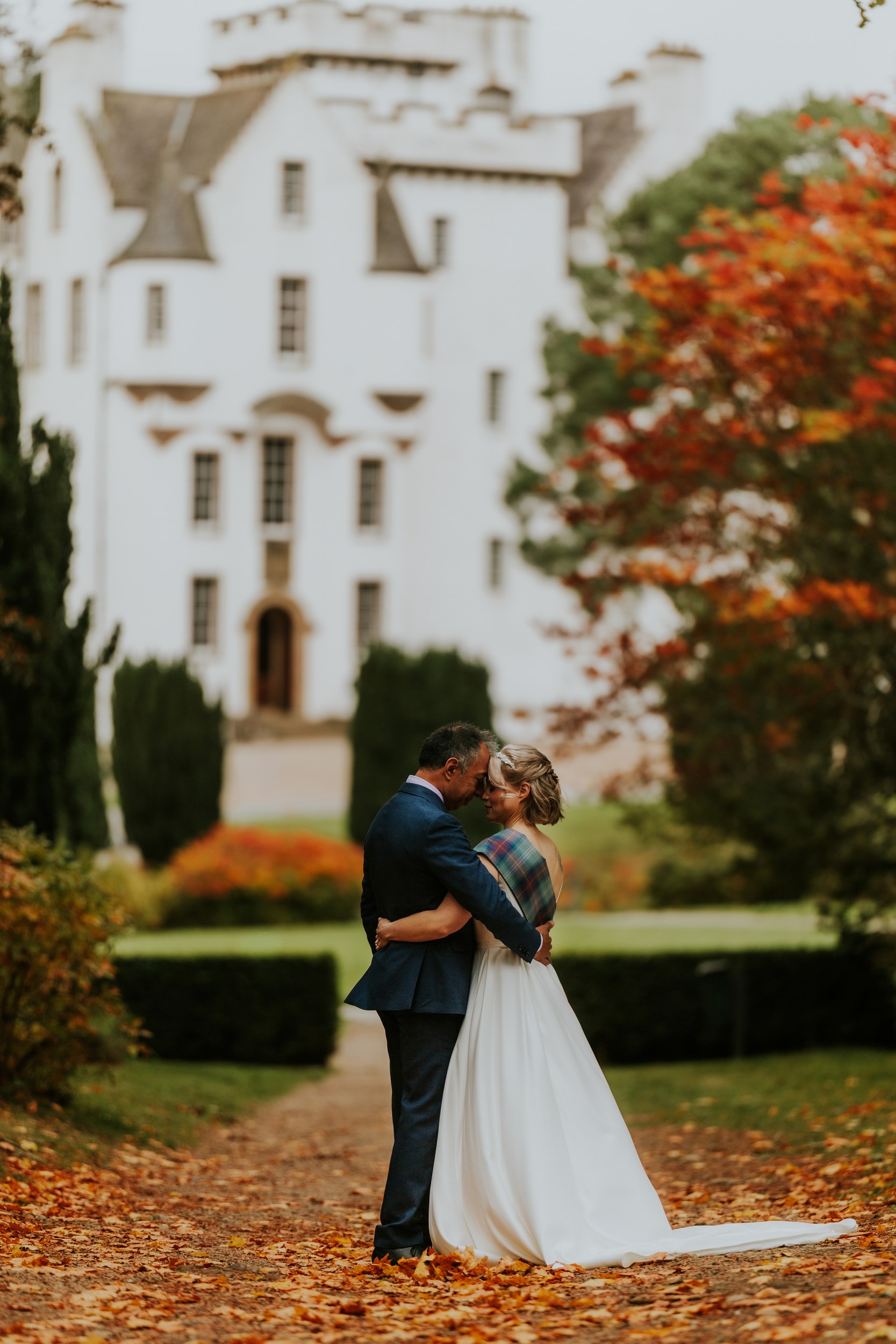 Bride and groom (Lizanne & Jitin) embrace on a leaf-covered path with Blair Castle blurred behind them.