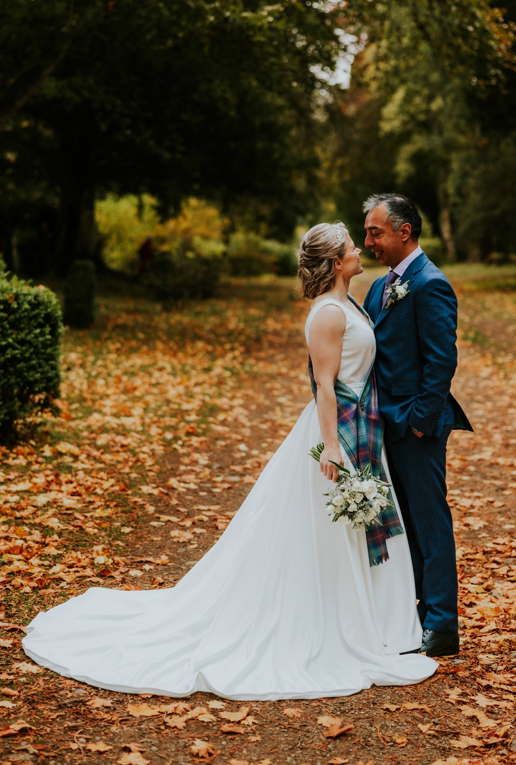 Bride and groom smiling on an autumn path; bride in white dress with tartan sash holds bouquet, groom in blue suit.