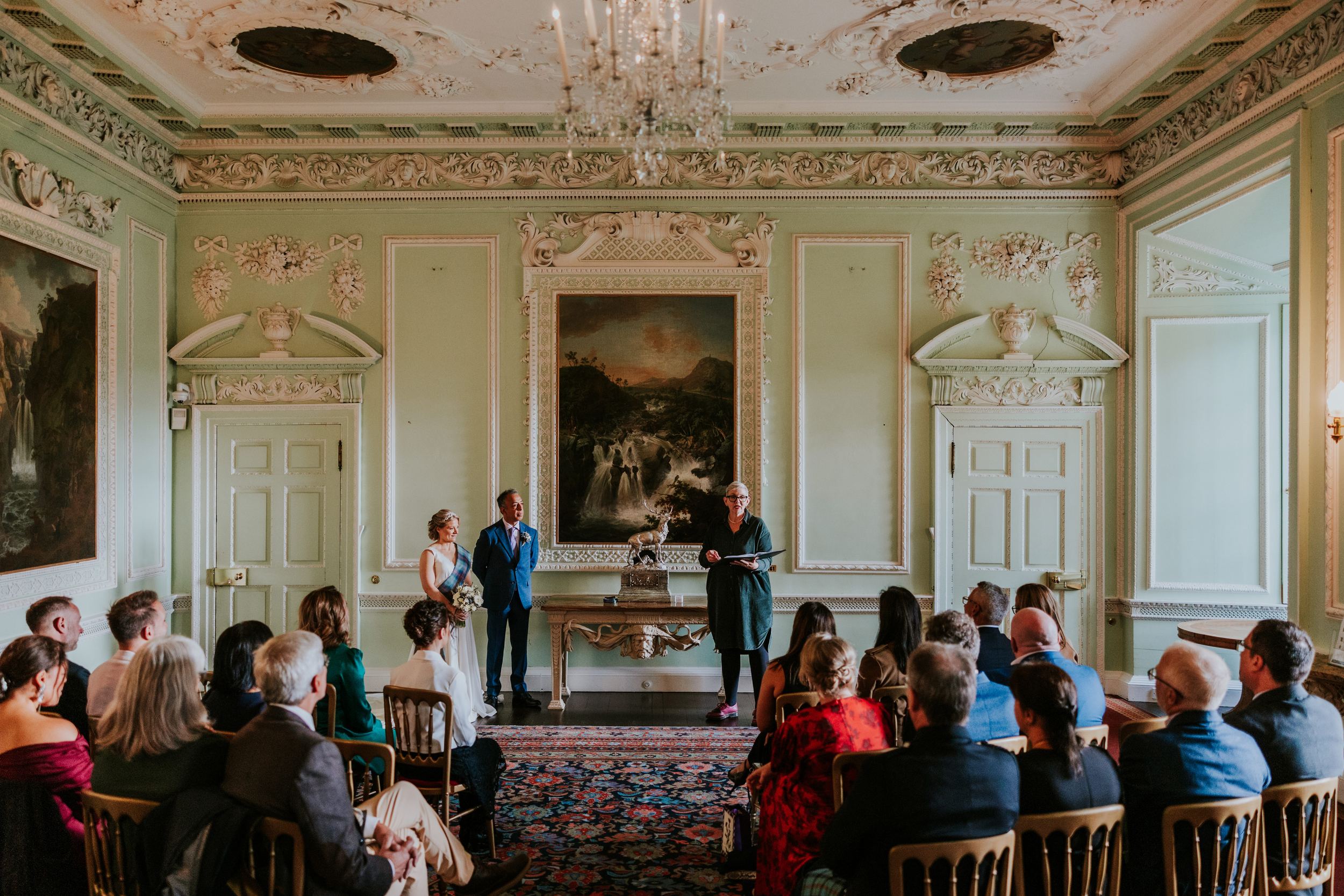 Bride and groom stand with the registrar in an ornate pale-green castle room as guests watch their civil ceremony.