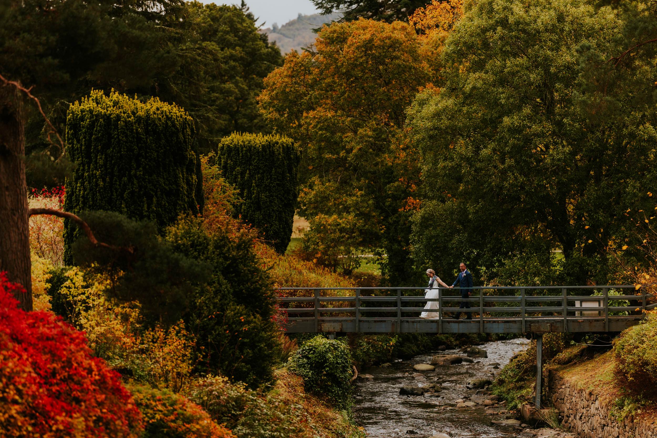 Bride and groom walk hand-in-hand across a bridge over a stream amid autumn trees on estate grounds.