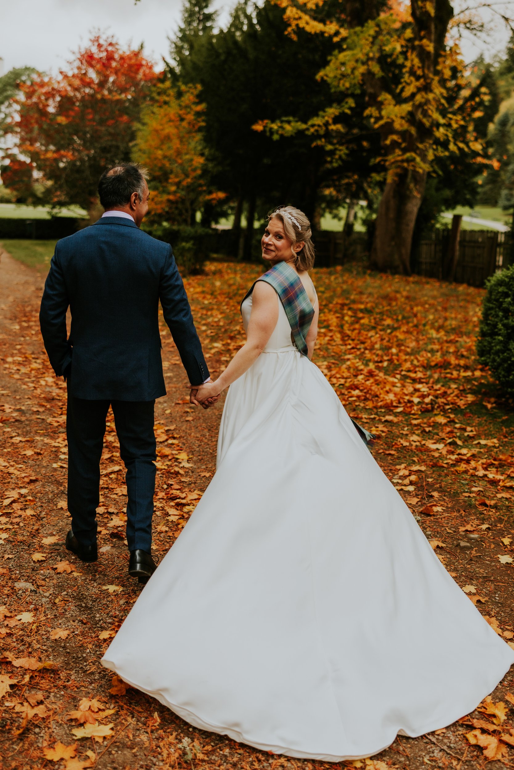 Bride and groom walk hand-in-hand on a leaf-strewn path; bride in white dress with tartan sash looks back and smiles.