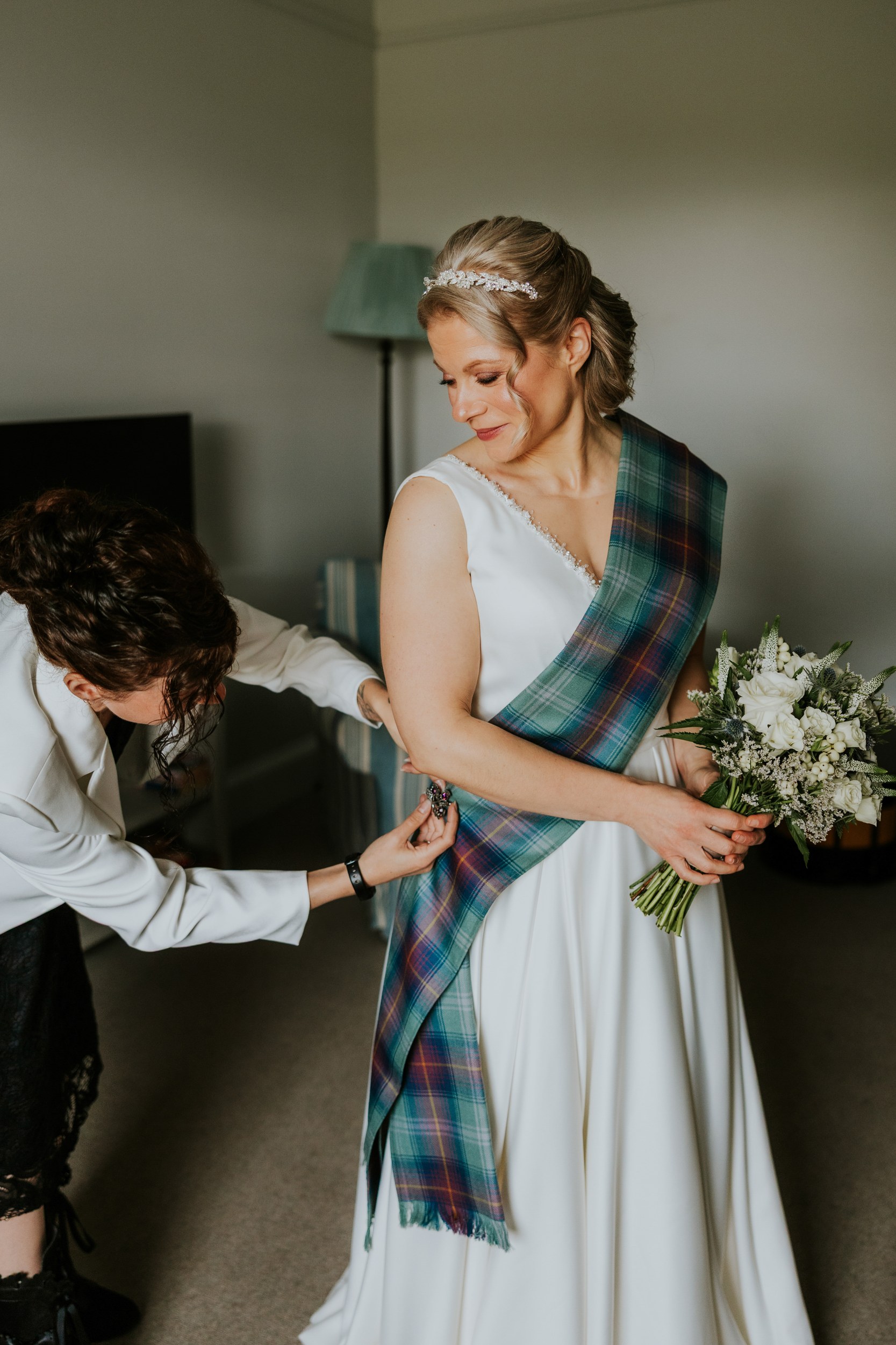 Bride having a green tartan sash fastened over her white dress while holding a white bouquet.