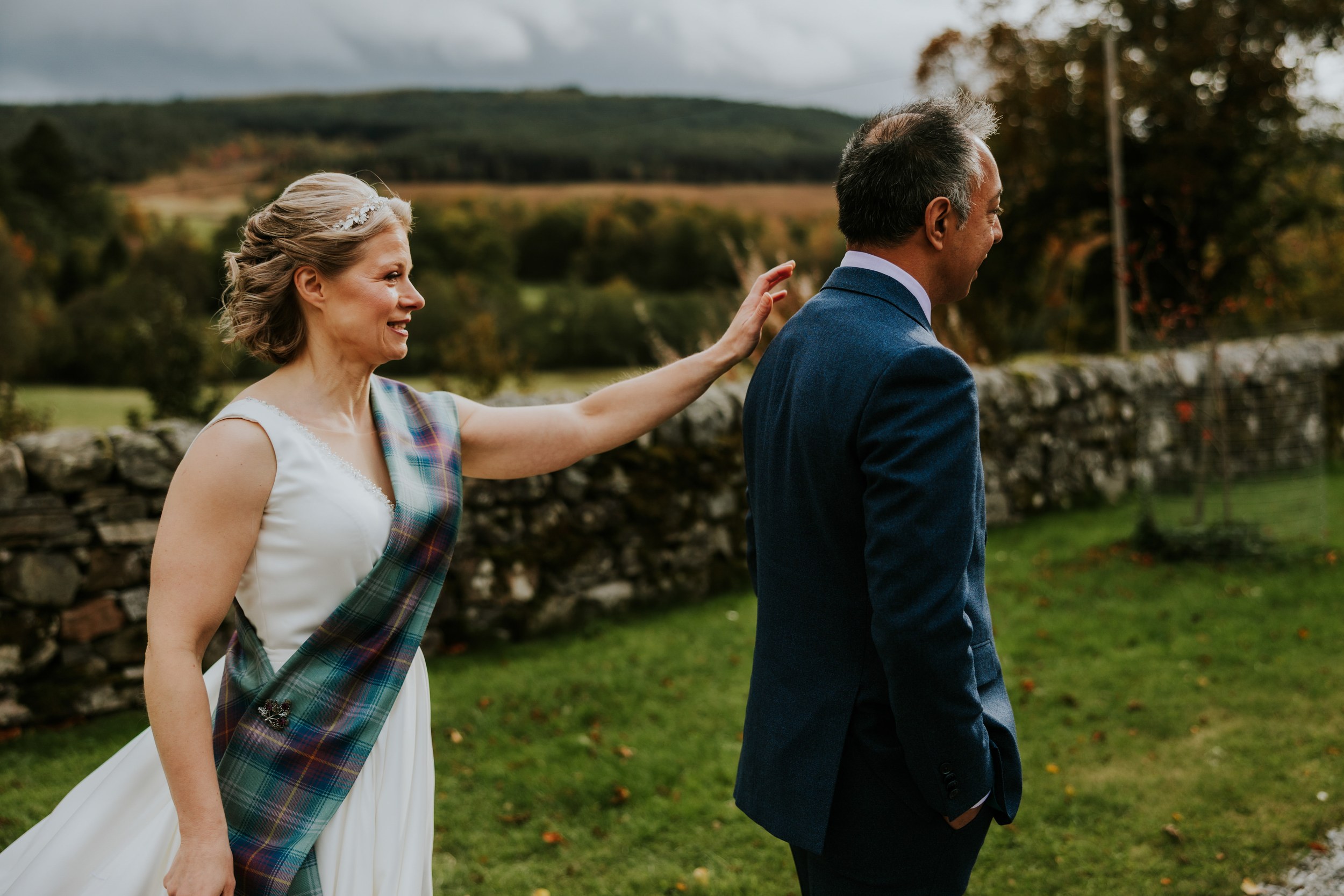 Bride in white dress with tartan sash touches groom's shoulder outdoors by a stone wall and rolling hills.