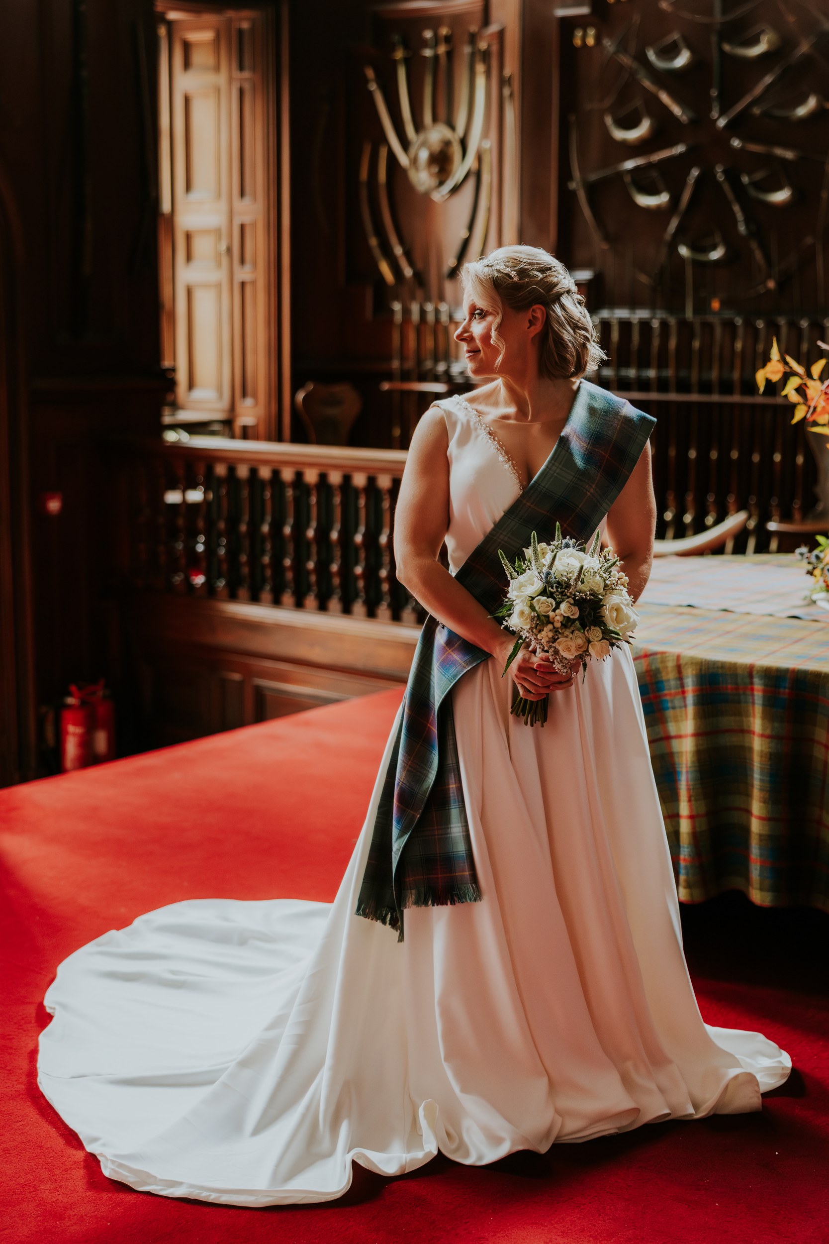 Bride in white dress with tartan sash holding bouquet on red carpet in wood‑panelled castle room.