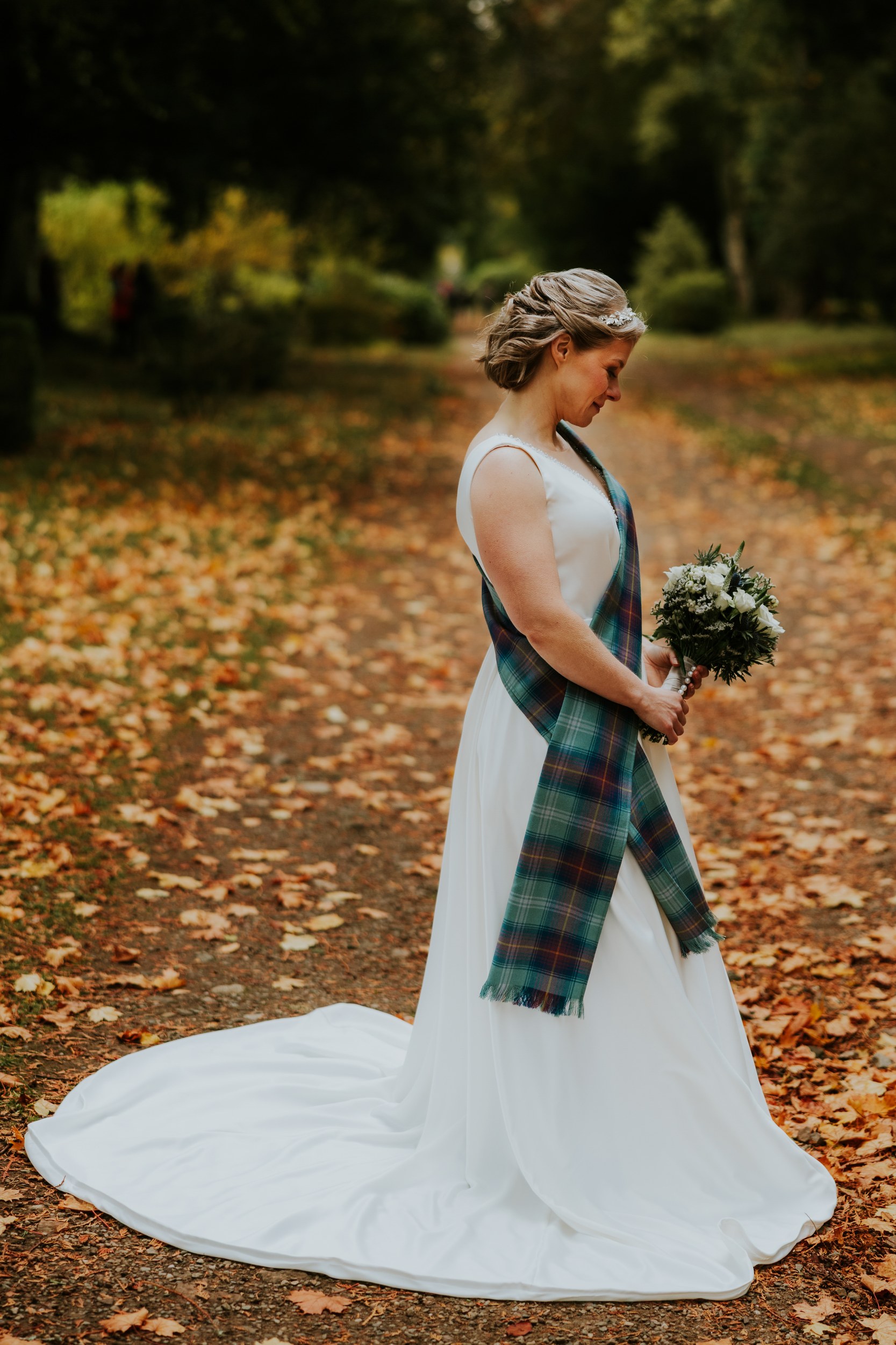 Bride in white dress with tartan sash holding a bouquet on an autumn leaf-strewn path, looking down.