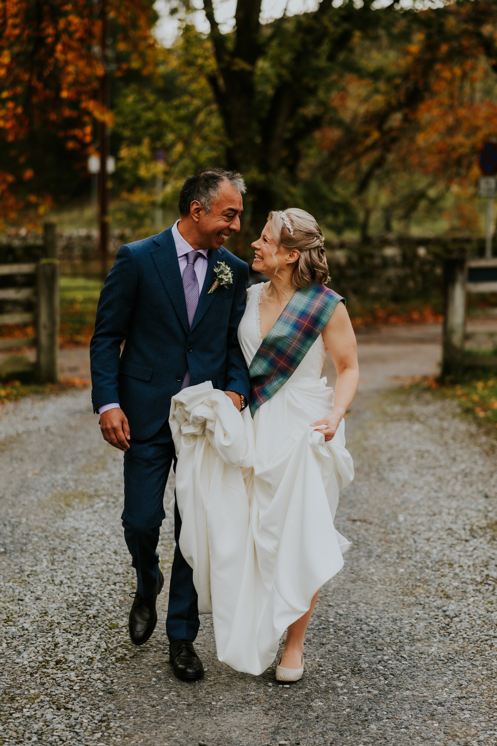 Bride in white dress with tartan sash and groom in blue suit walk smiling on a gravel path with autumn trees behind.
