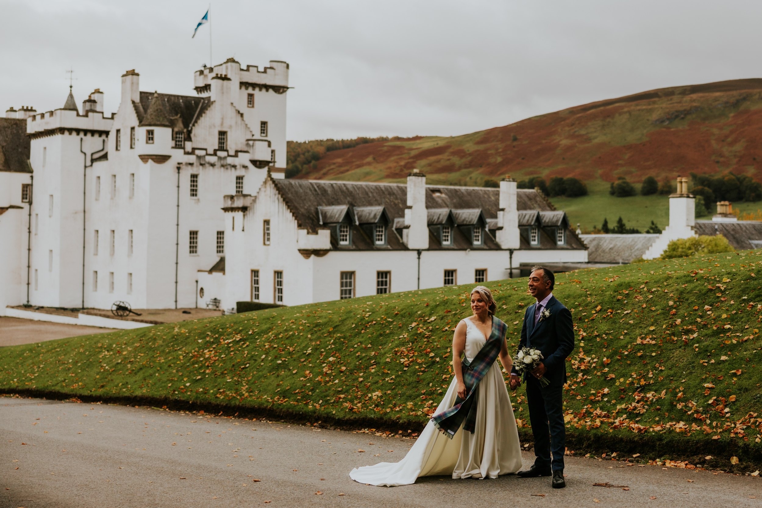 Bride in white dress with tartan sash and groom in suit walk by Blair Castle with autumn hills behind.