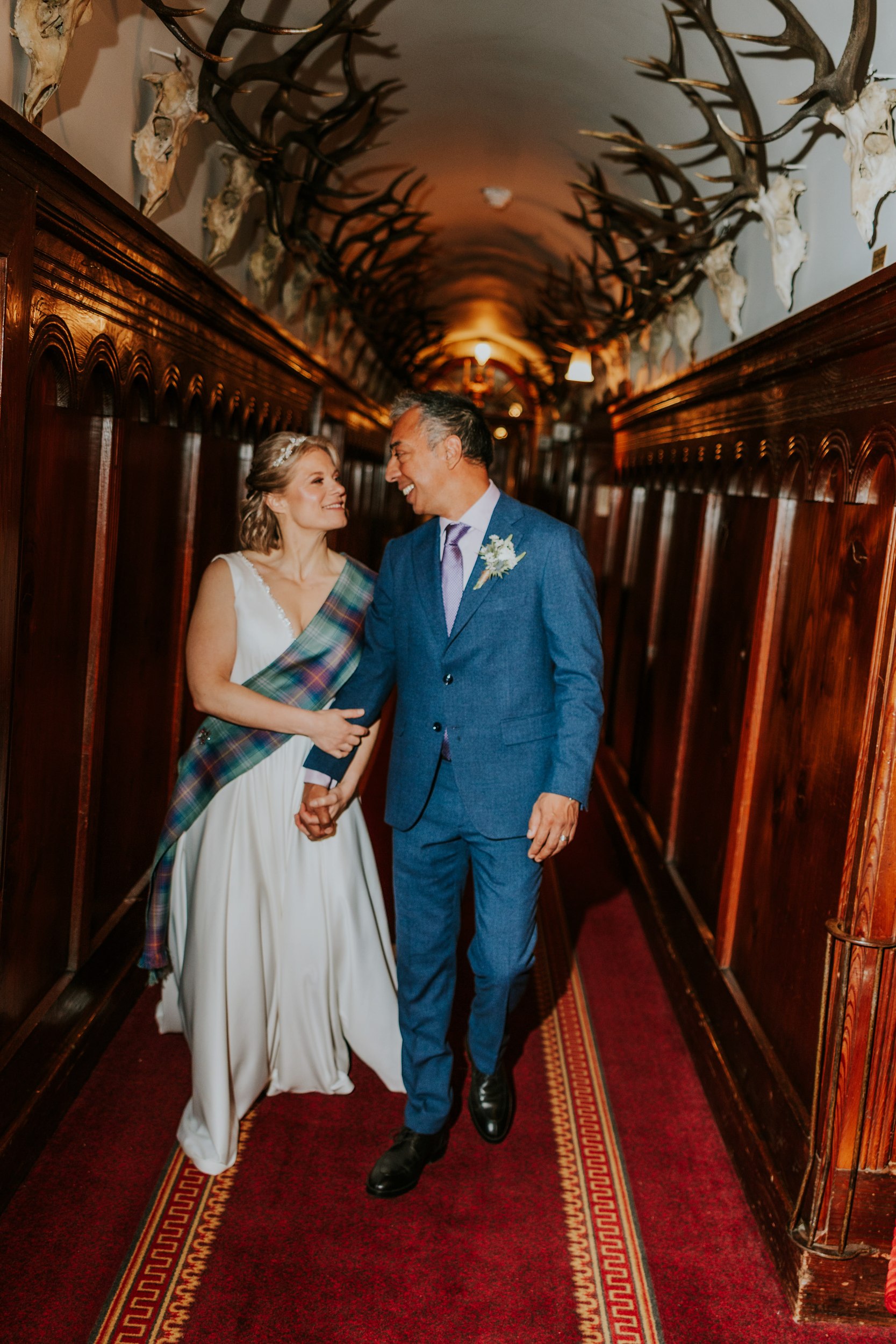 Bride in white dress with tartan sash and groom in blue suit holding hands in a wood‑panelled corridor lined with mounted stag antlers.