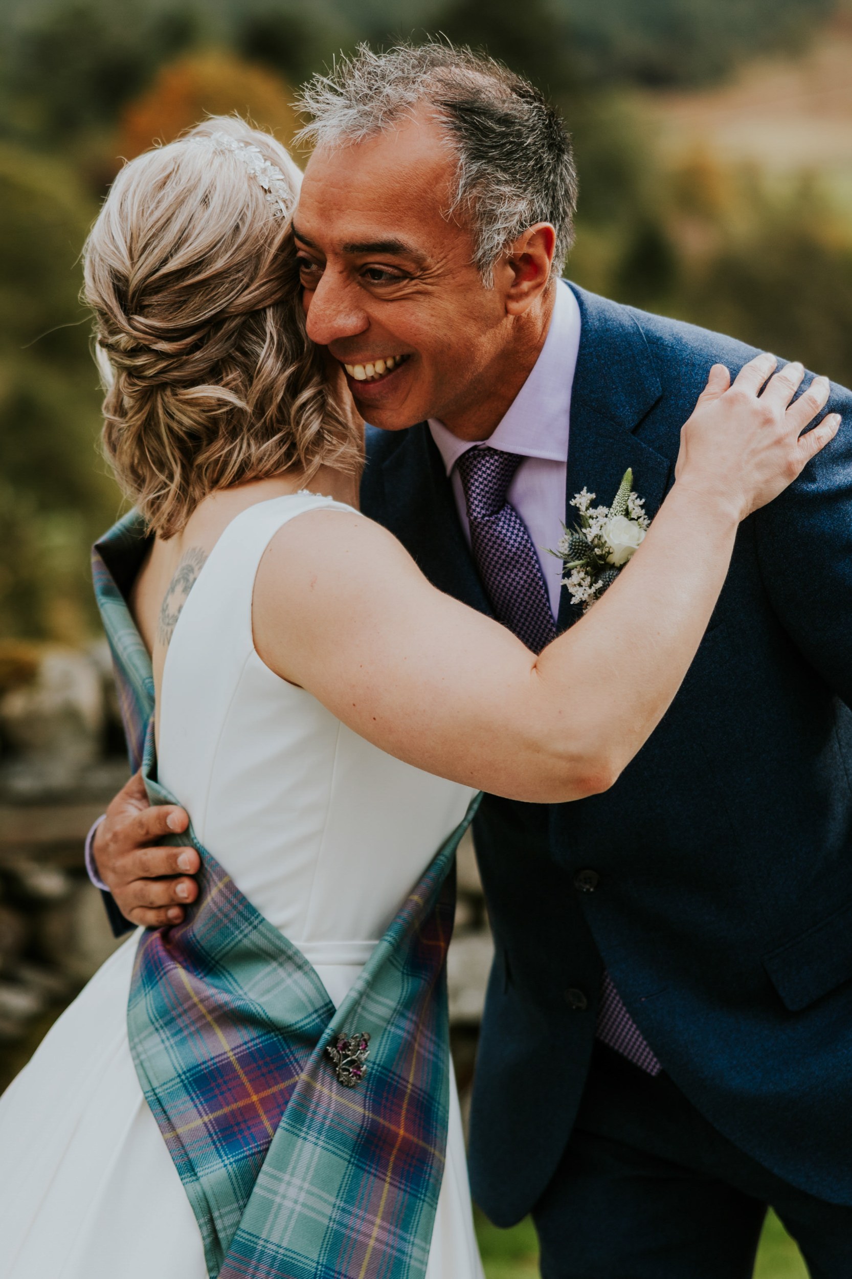 Bride in white dress with tartan sash hugs smiling groom in blue suit and purple tie outdoors.