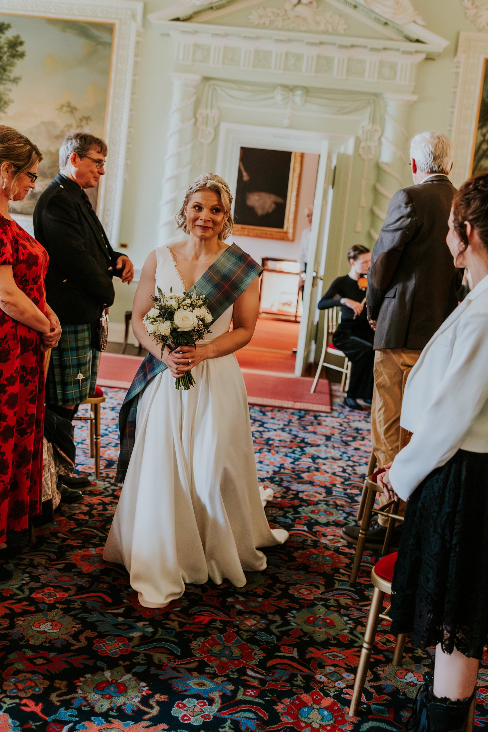 Bride in white gown with tartan sash holding bouquet walking down aisle in ornate room as guests stand
