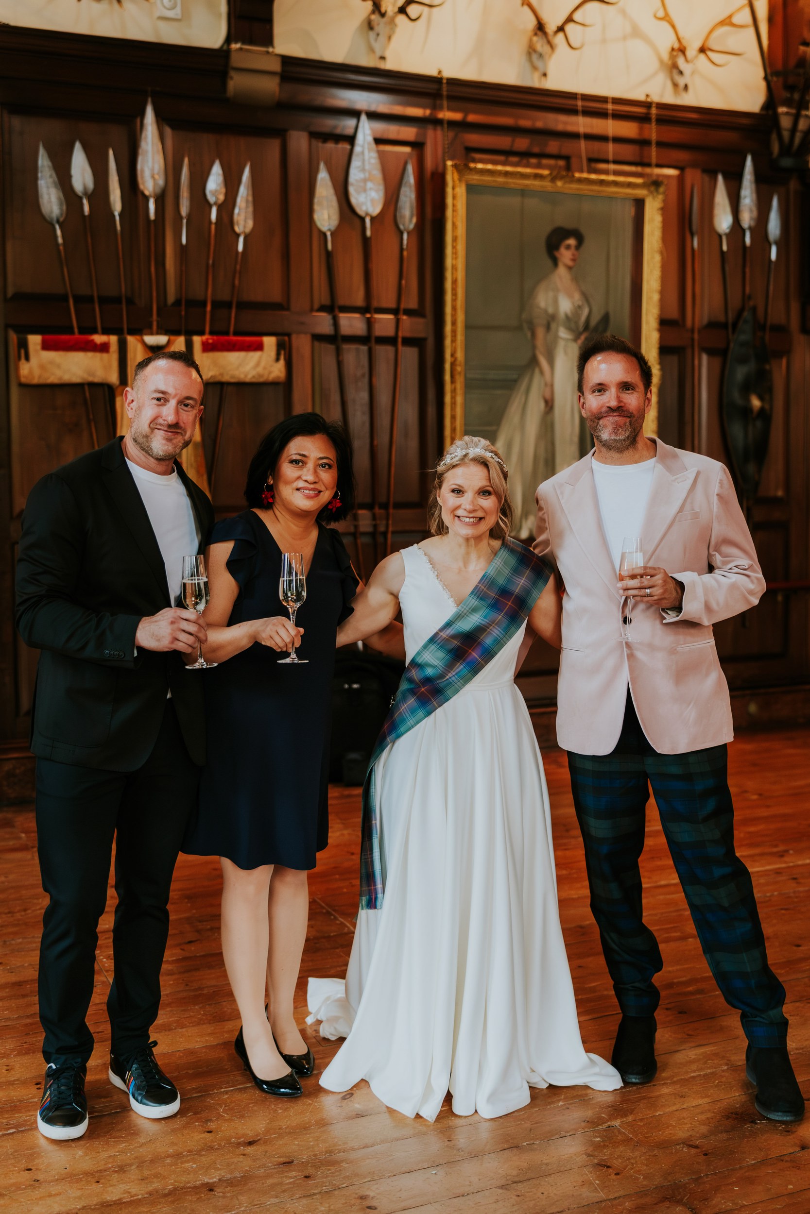 Bride in white with tartan sash and three guests holding champagne in a wood‑panelled castle room with portrait and mounted spears.