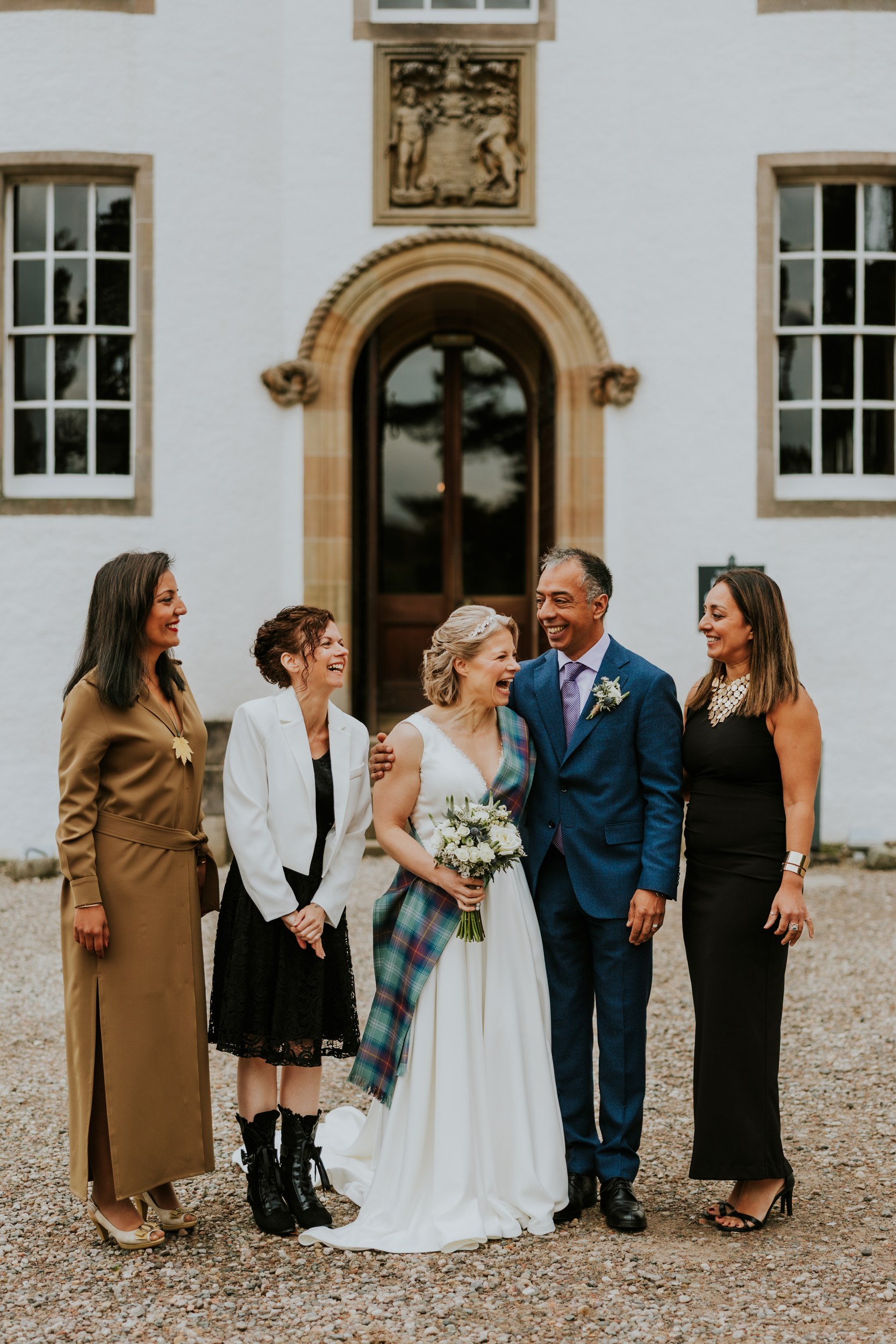 Bride Lizanne in white with tartan sash and groom Jitin in a blue suit laugh with three guests outside the castle entrance.