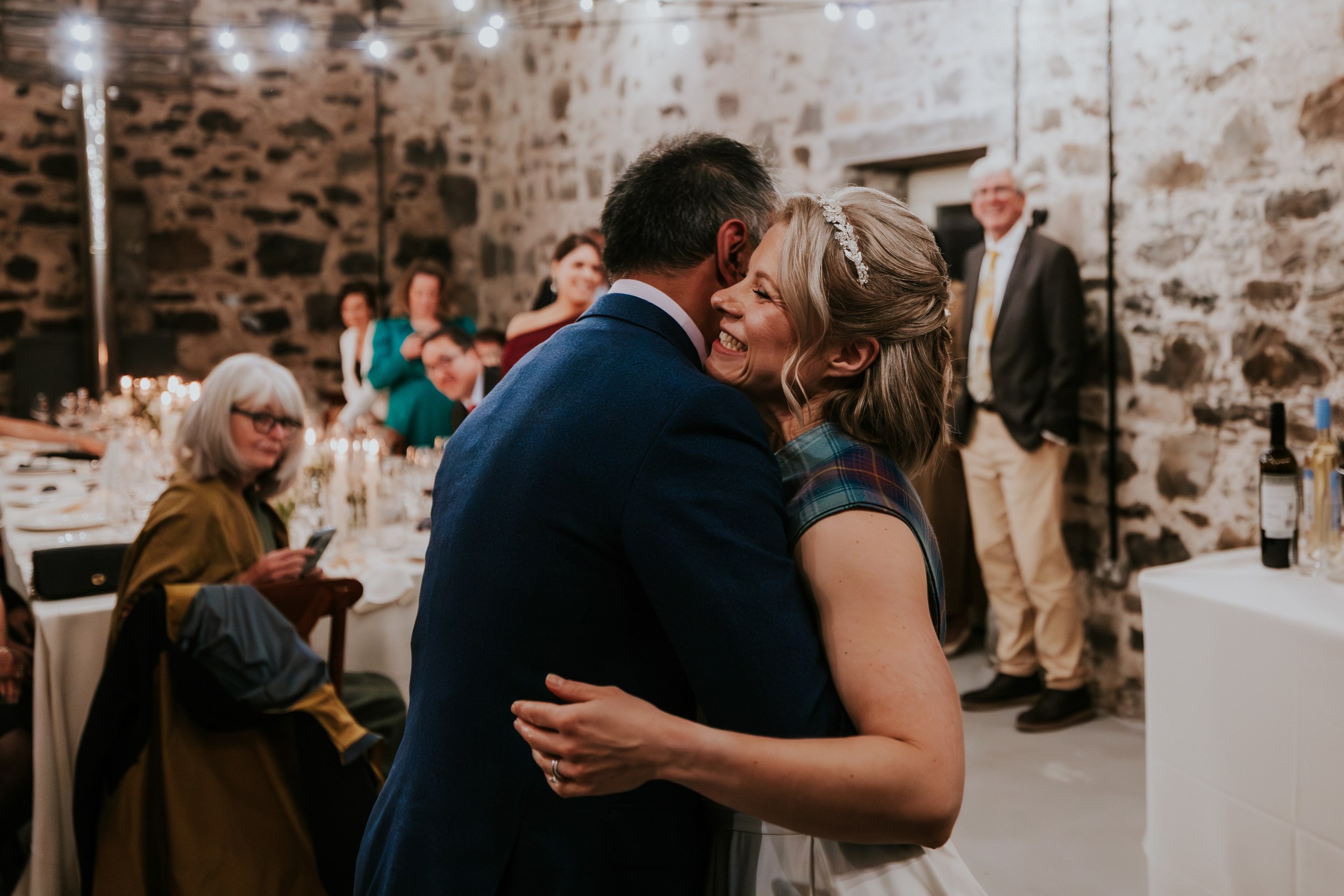 Bride smiling as she hugs a man at a stone-walled reception barn, guests and candlelit tables behind.