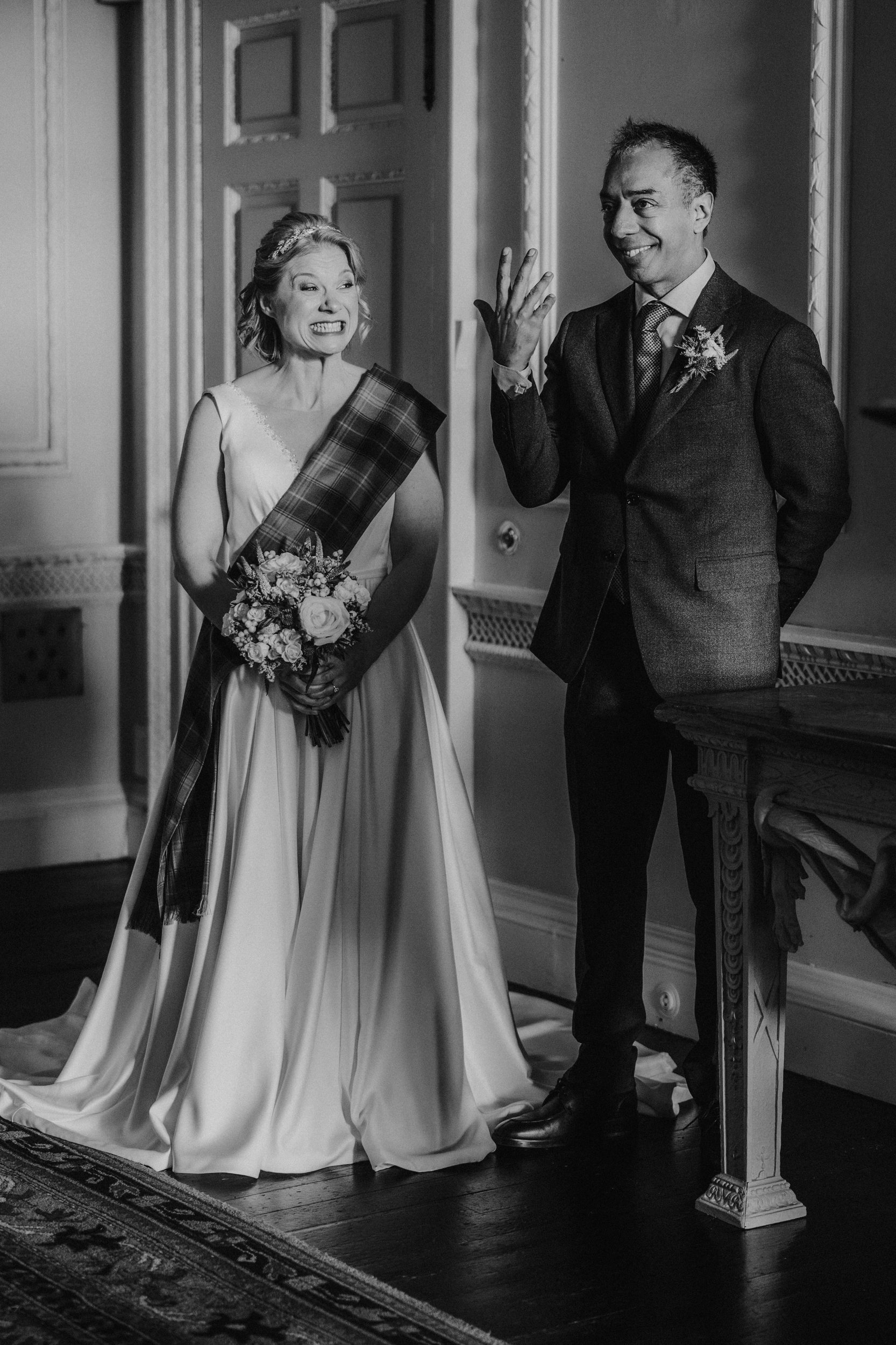 Bride with bouquet and tartan sash beside groom who smiles and raises his hand inside a castle ceremony room.