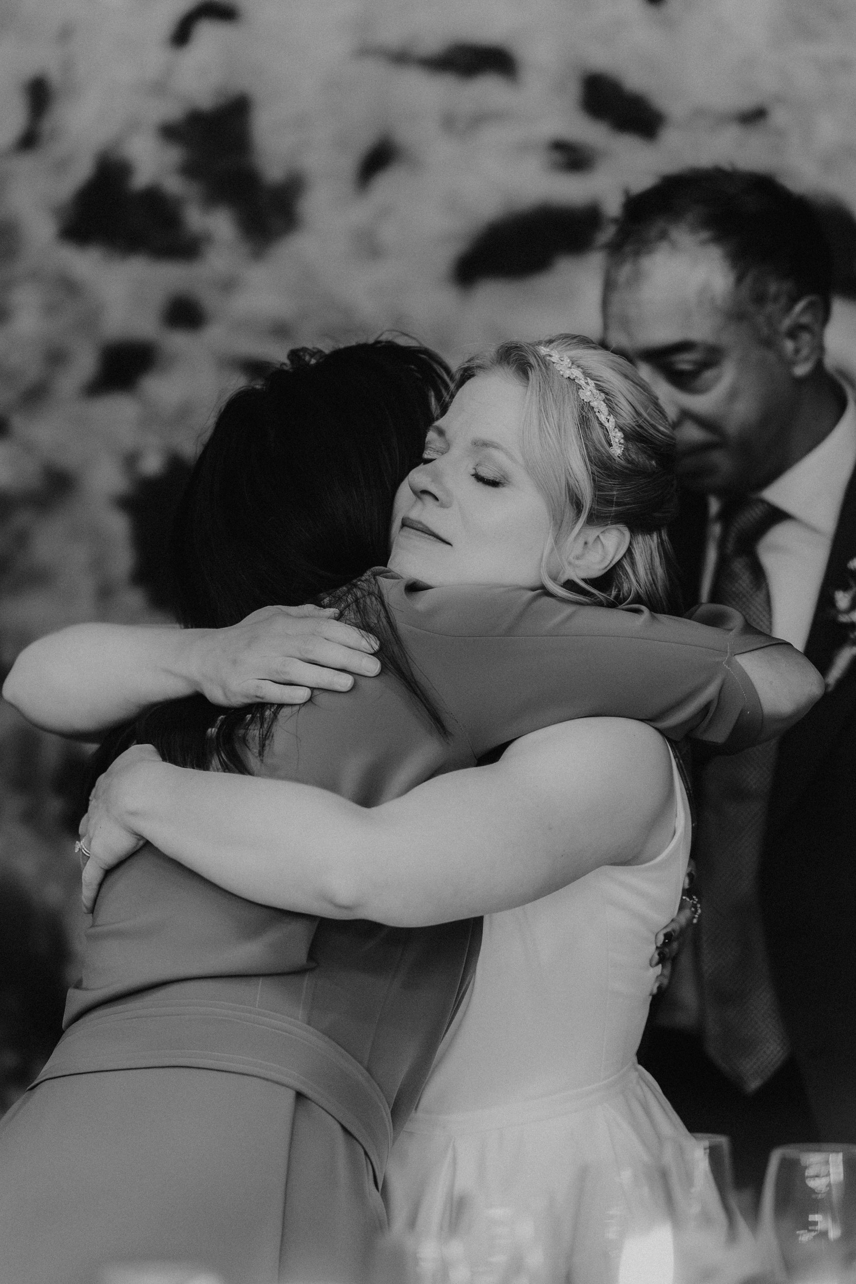Bride with hairpiece closes her eyes while hugging a guest, a man in a suit stands behind them.