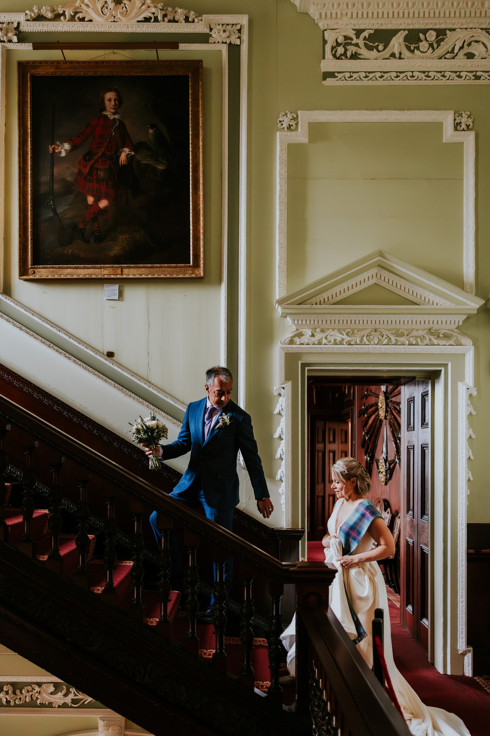 Bride with tartan sash and man in blue suit descending ornate staircase inside a castle with a large portrait on the wall.