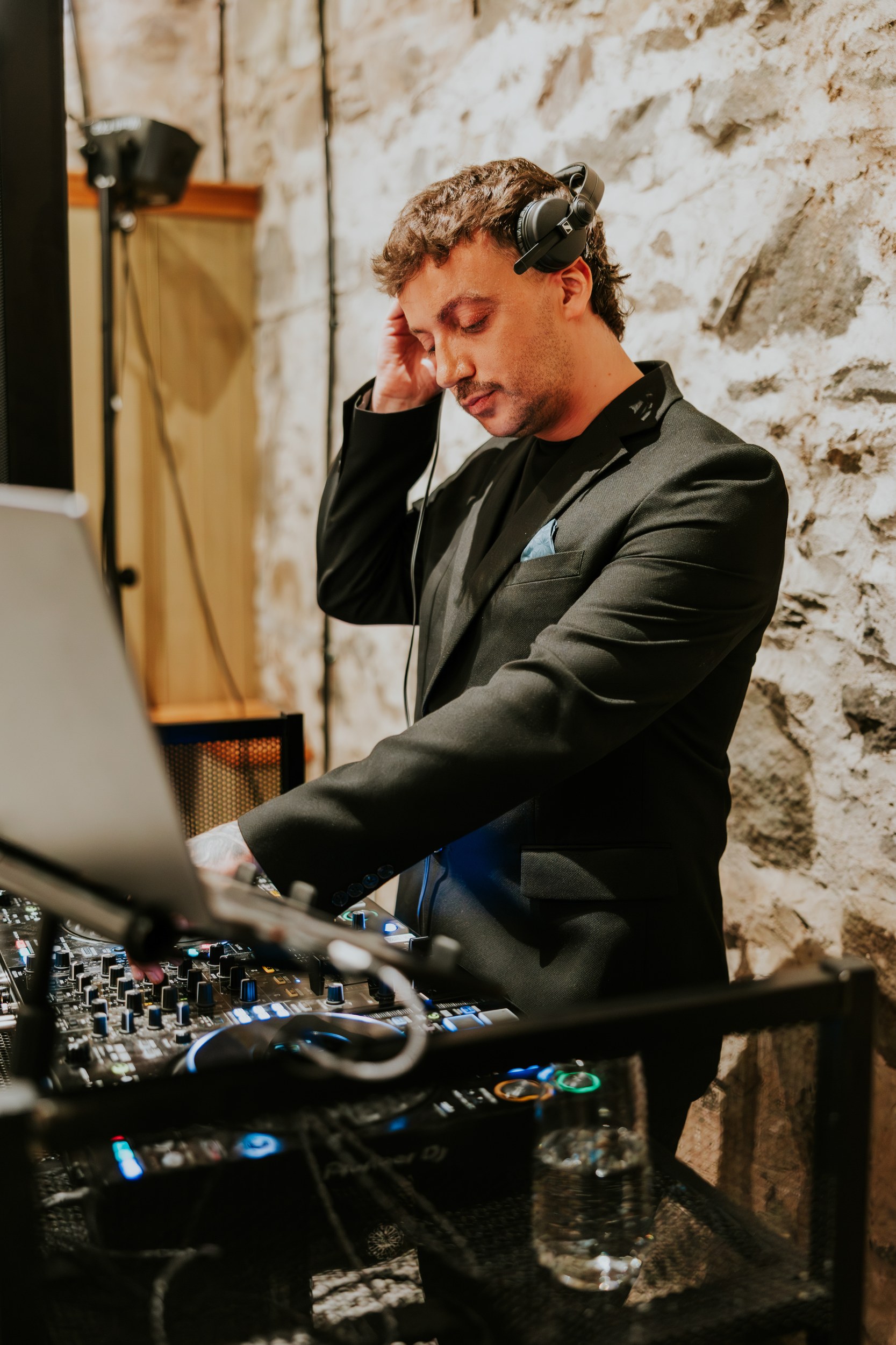 DJ in a black suit with headphones adjusting a mixer against a stone wall in the reception space.