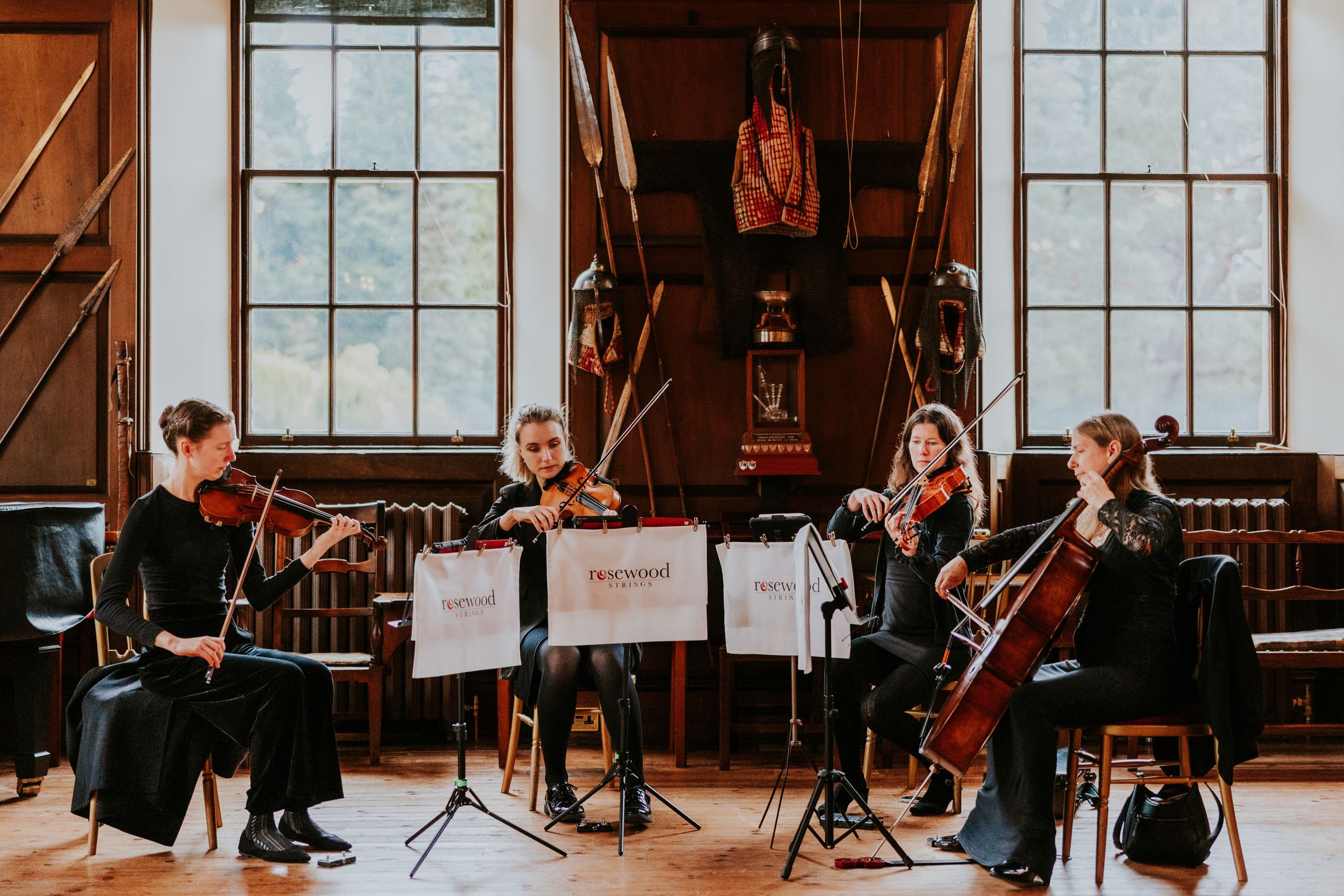 Four-piece string quartet performing in a wood‑panelled room with tall windows and displayed armour.