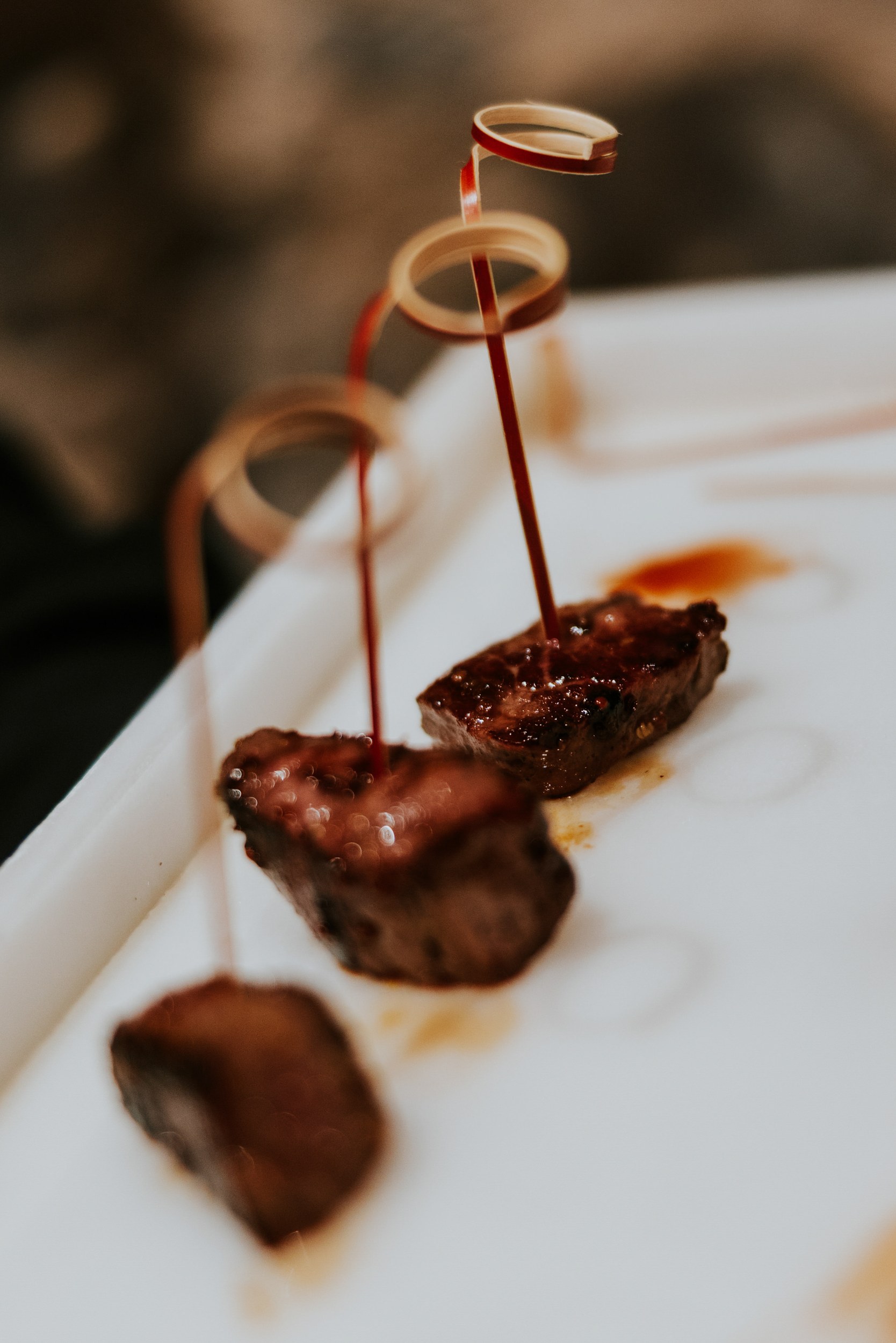 Glazed meat canapés on skewers with curled red cocktail picks on a white tray.