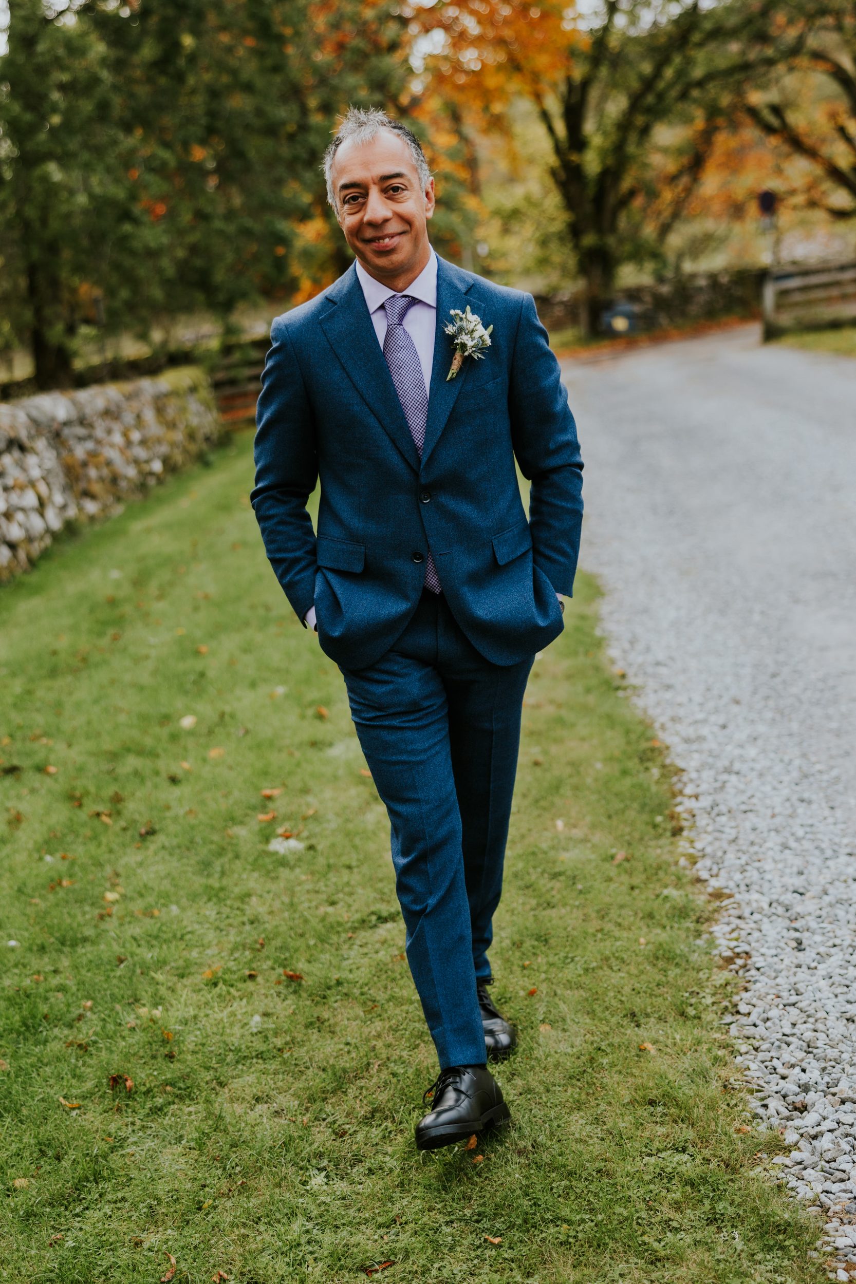 Groom Jitin in a blue suit walking on grass beside a gravel drive with autumn trees behind.