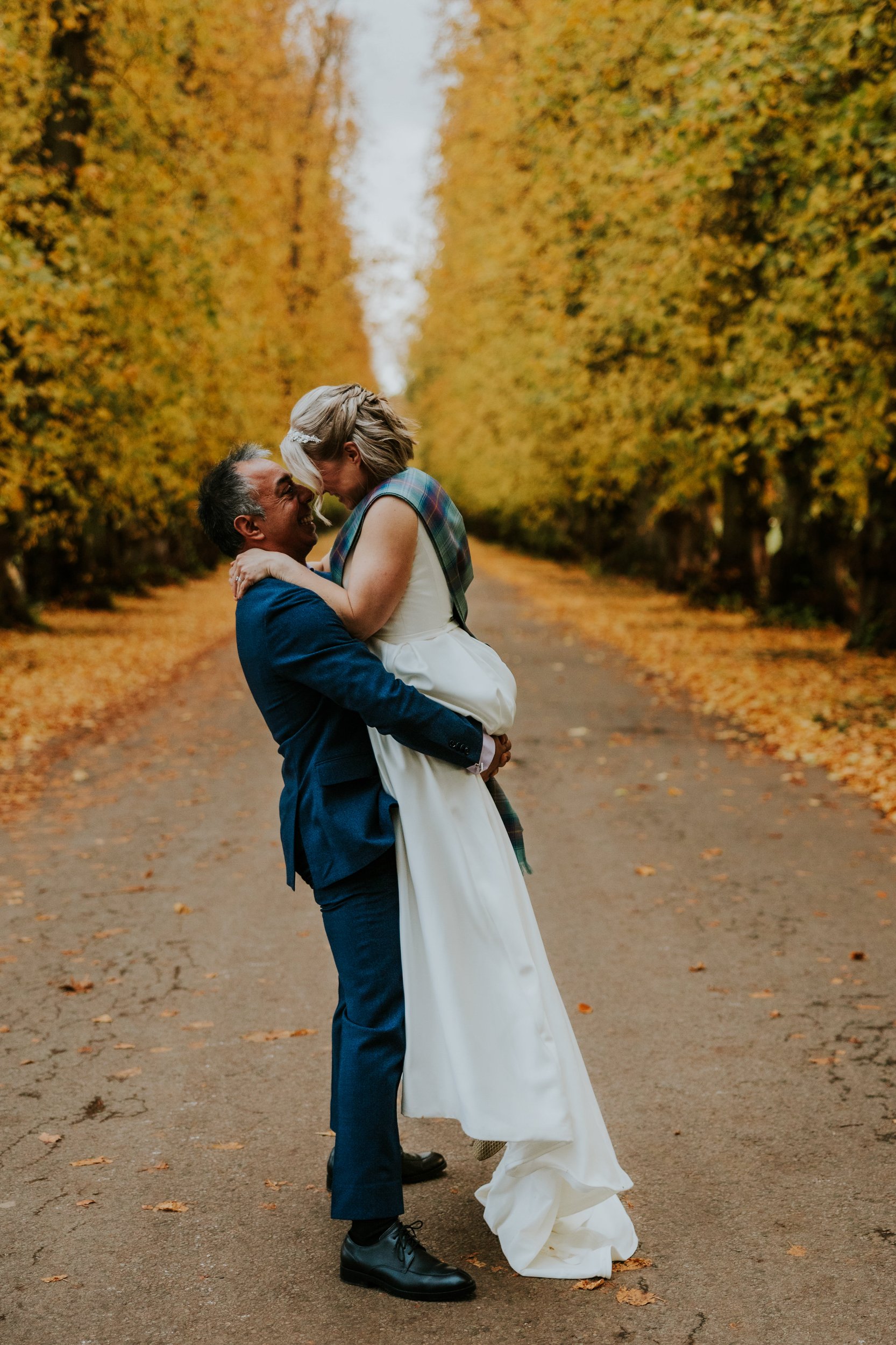 Groom lifts bride on a tree-lined, leaf-strewn avenue at Atholl Estate, Perthshire.