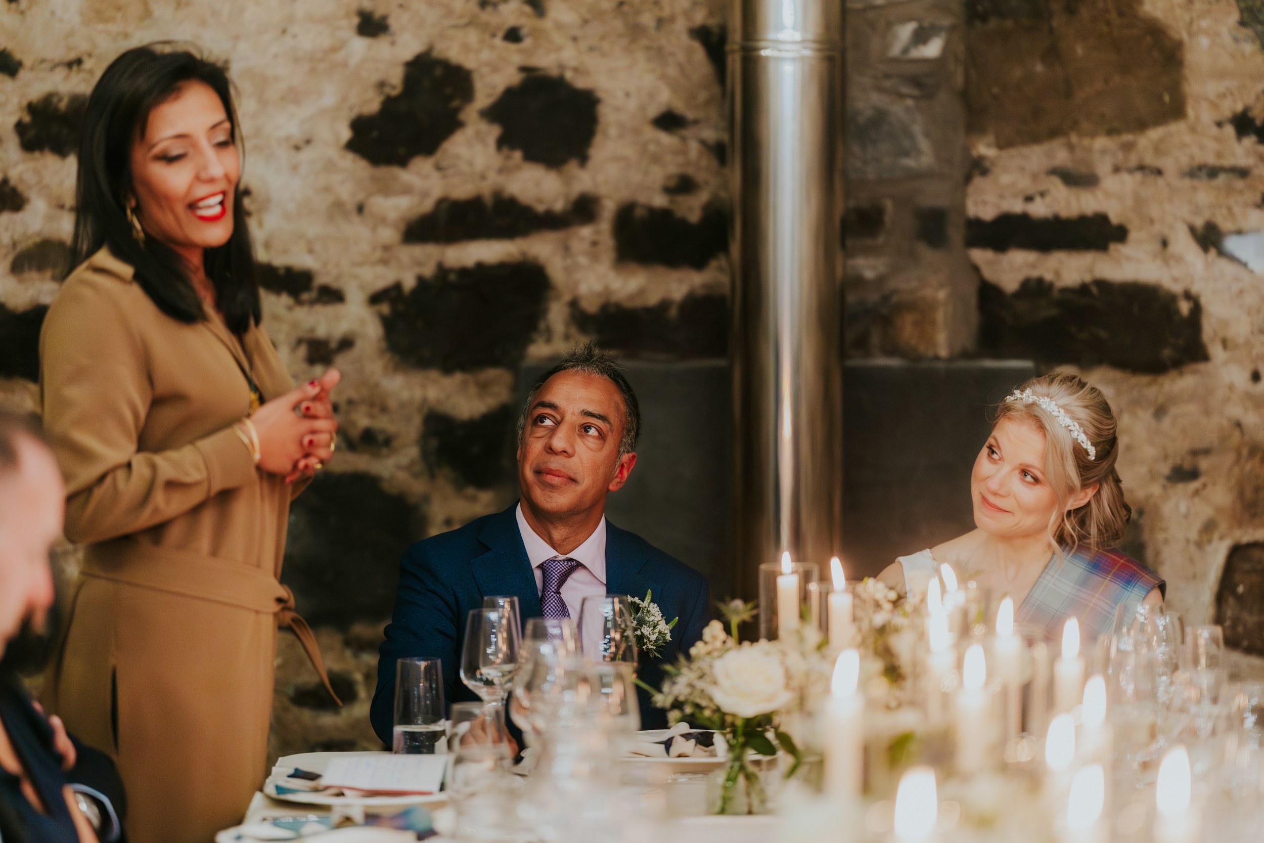 Guest speaking as bride Lizanne and groom Jitin listen at a candlelit head table in a stone-walled reception barn.