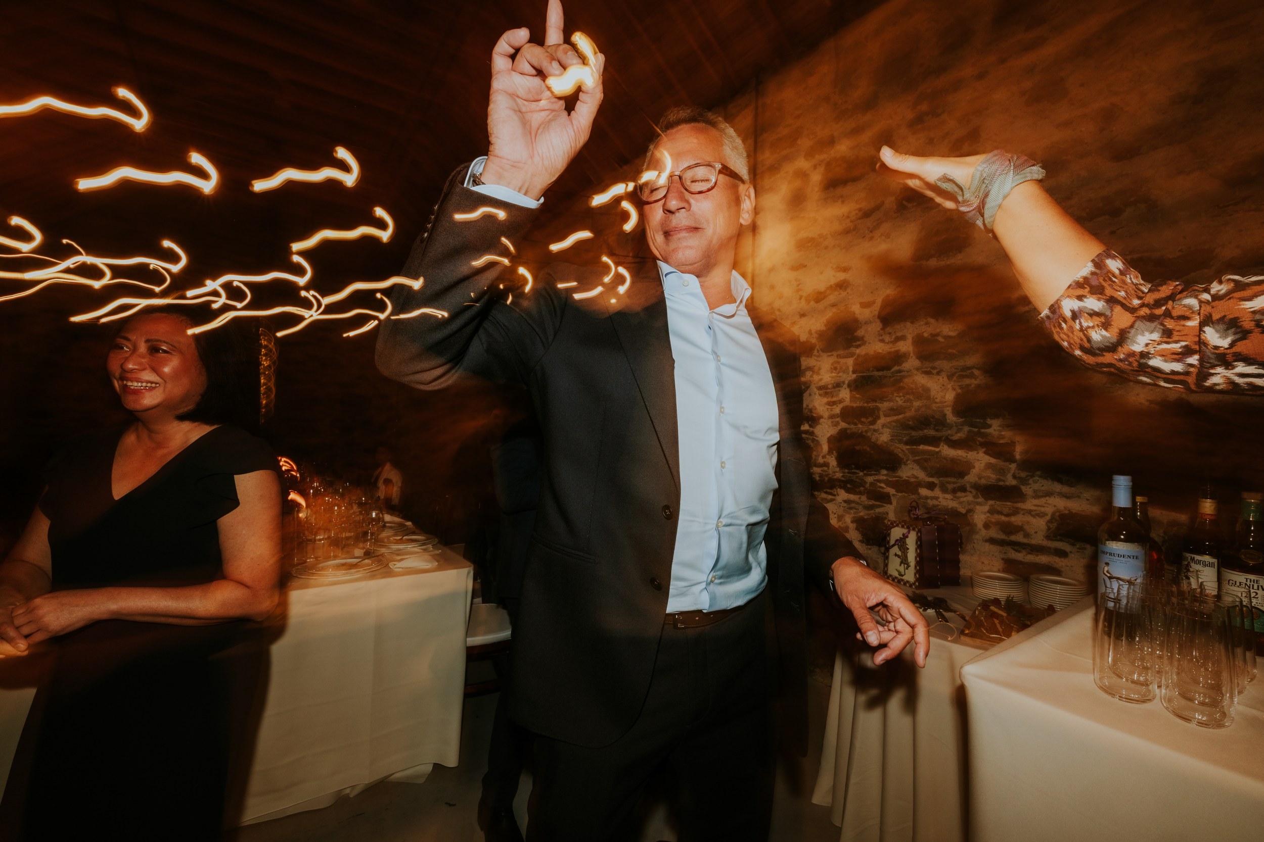 Guests dancing in the Clachan reception barn with golden light trails; man in a suit raises a finger and a woman smiles.