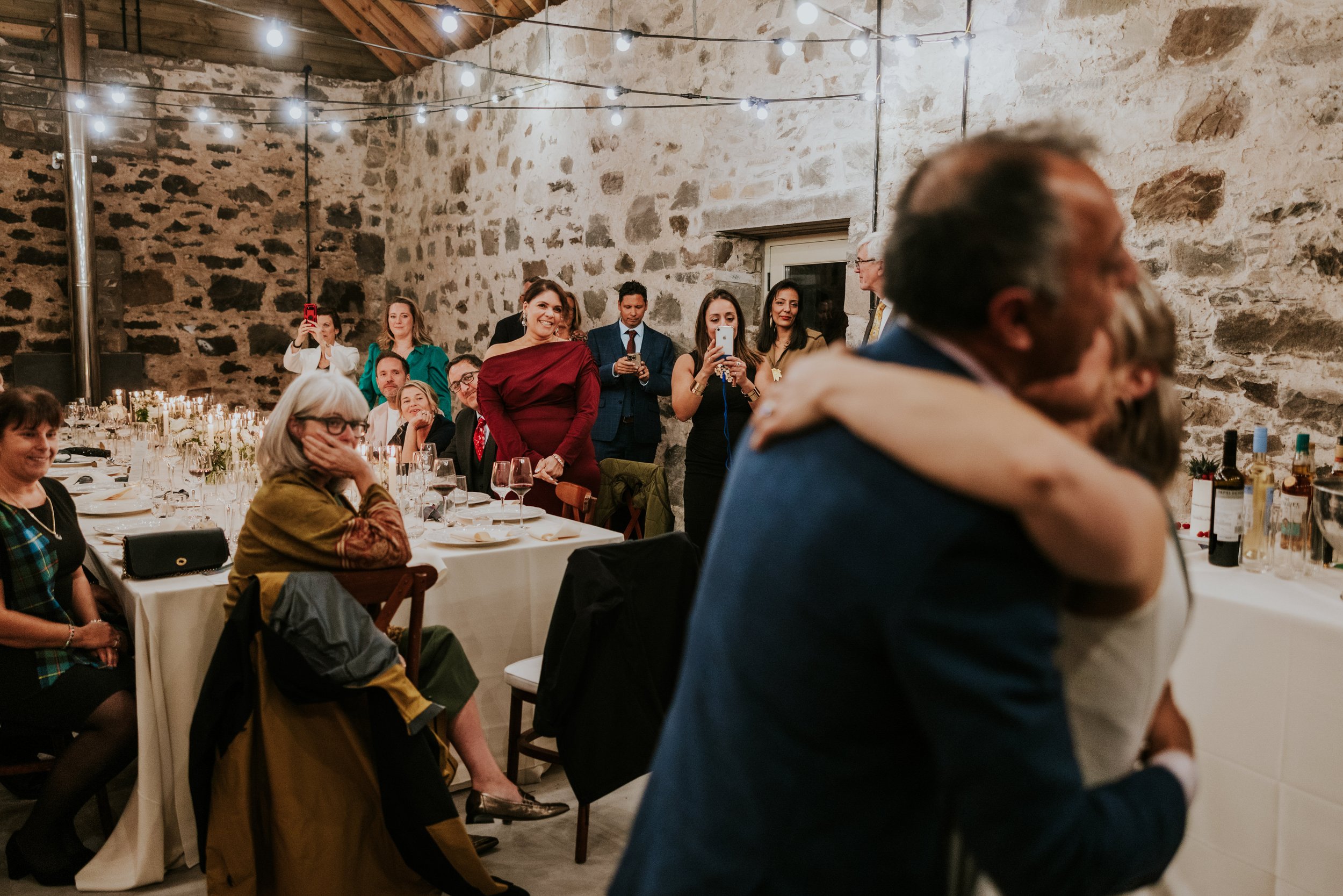Guests in a stone-walled barn watch and photograph a couple embracing in the foreground.