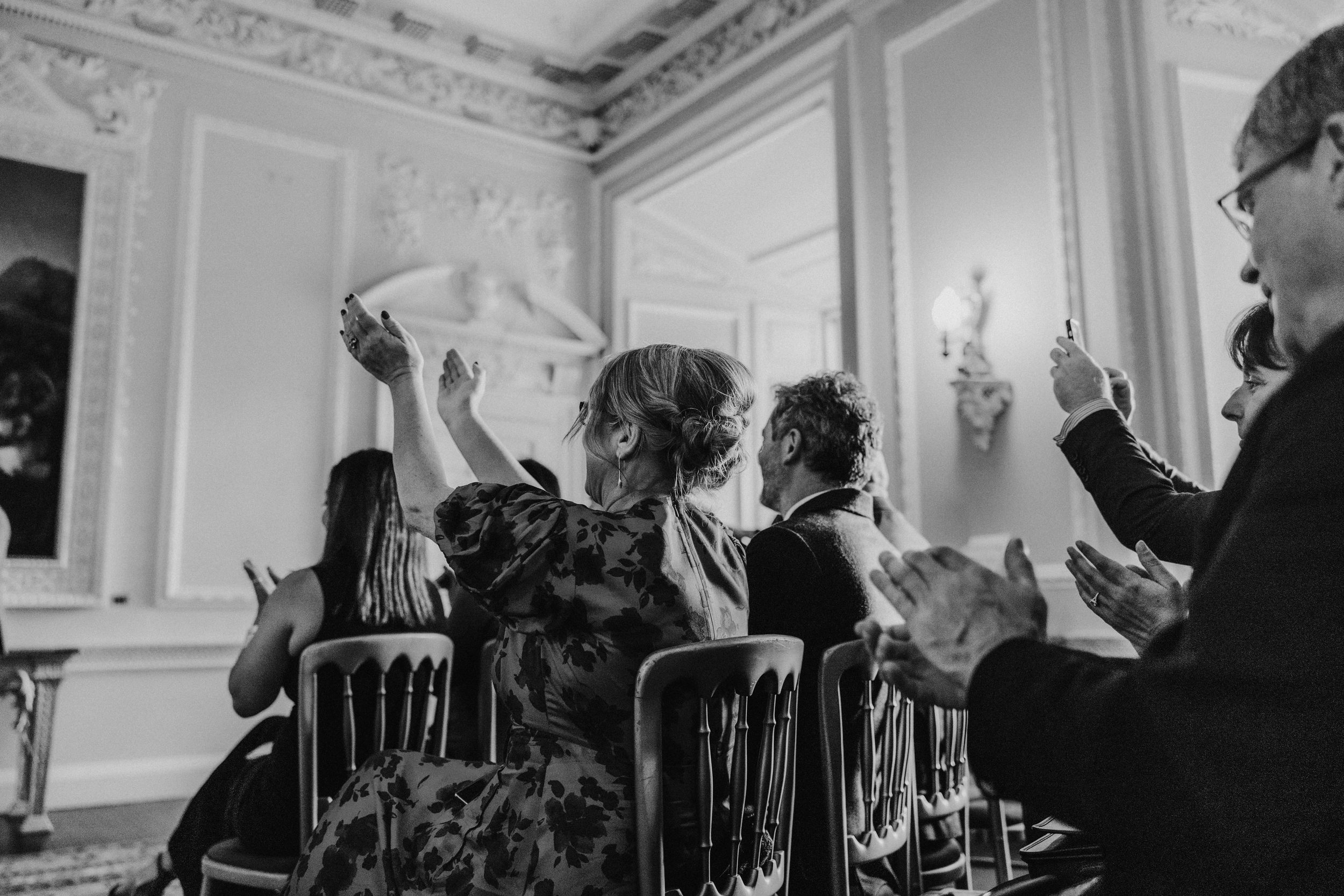 Guests in an ornate room applaud and raise their hands during the ceremony; one guest holds up a phone.
