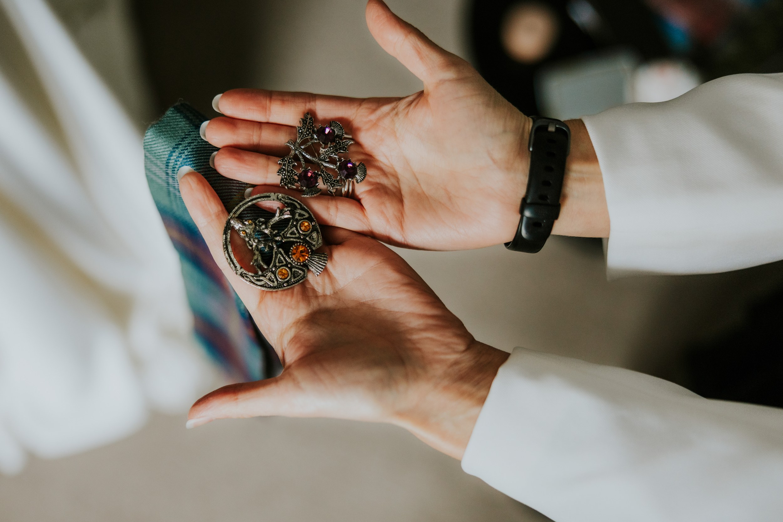Hands holding two vintage brooches: a round bird brooch with amber stones and a thistle brooch with purple gems, tartan tie nearby.