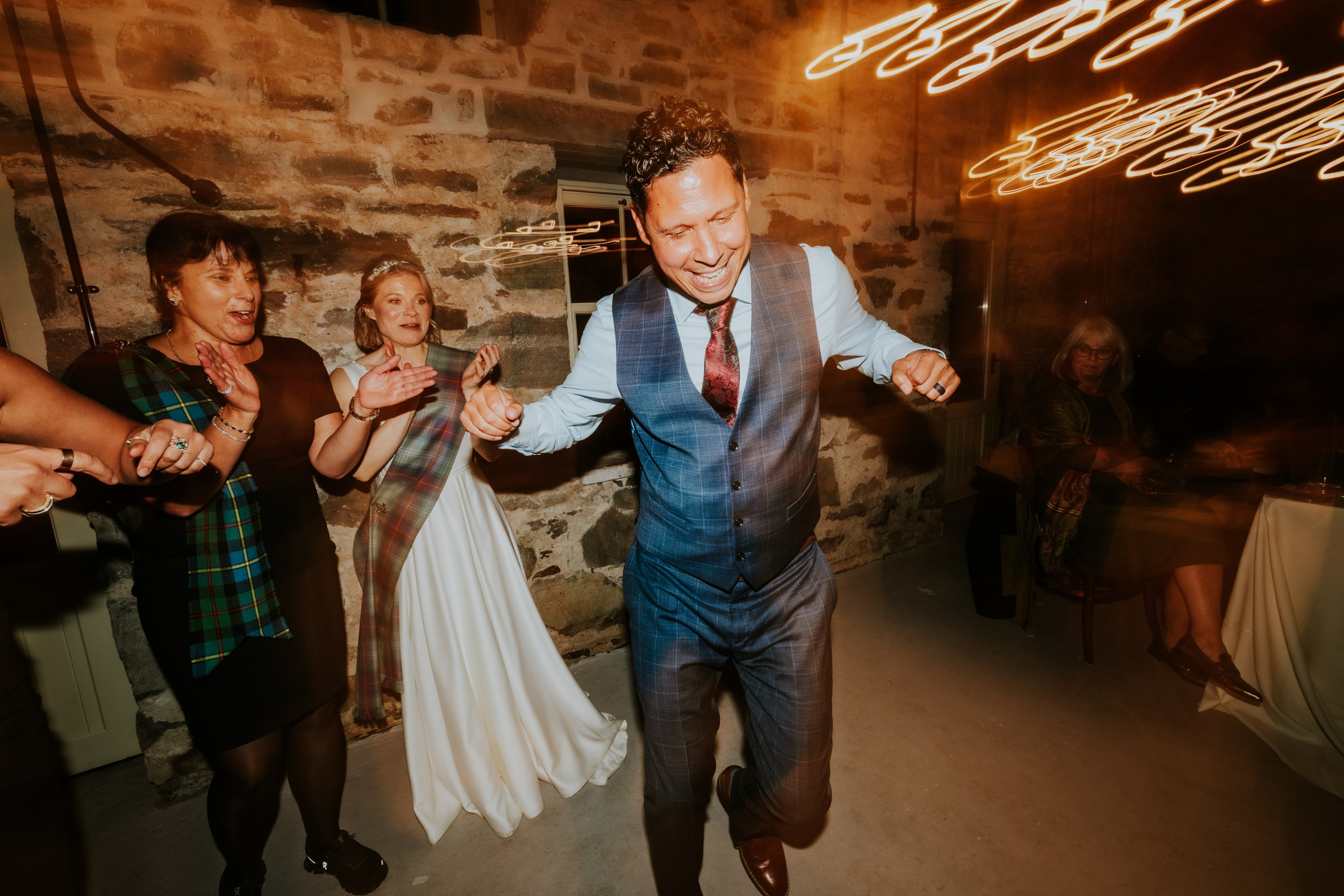 Lizanne and Jitin dancing in a stone-walled reception barn, bride with tartan sash and guests clapping under warm motion-lit lights.