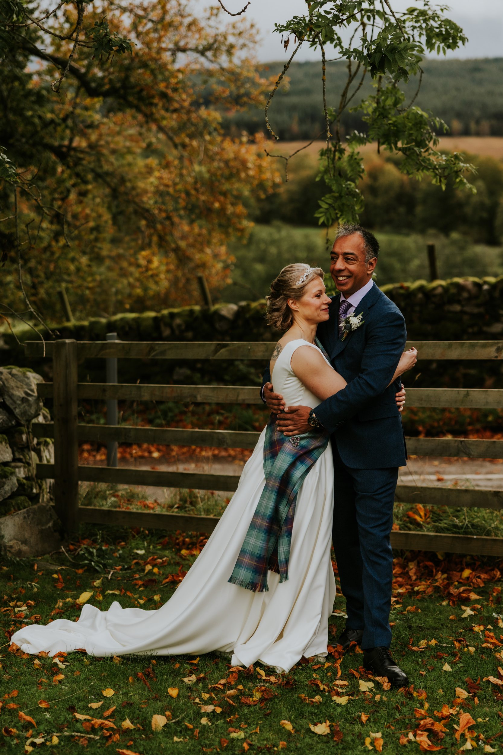 Lizanne and Jitin embrace by a wooden fence, bride in white dress with tartan sash, groom in a blue suit; autumn fields and stone wall behind.