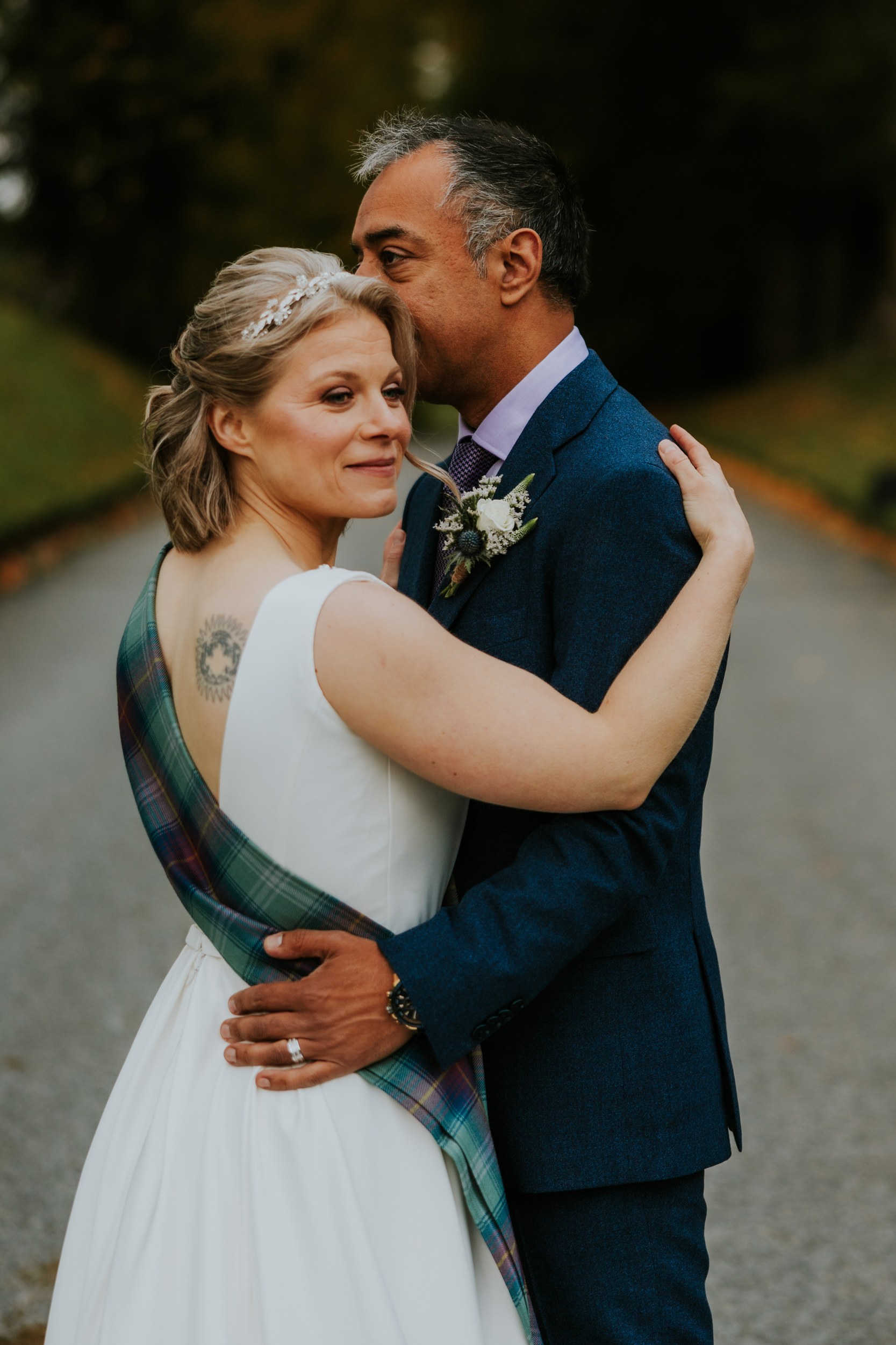 Lizanne and Jitin embrace on a tree-lined drive; bride in white dress with tartan sash, groom in blue suit kissing her head.