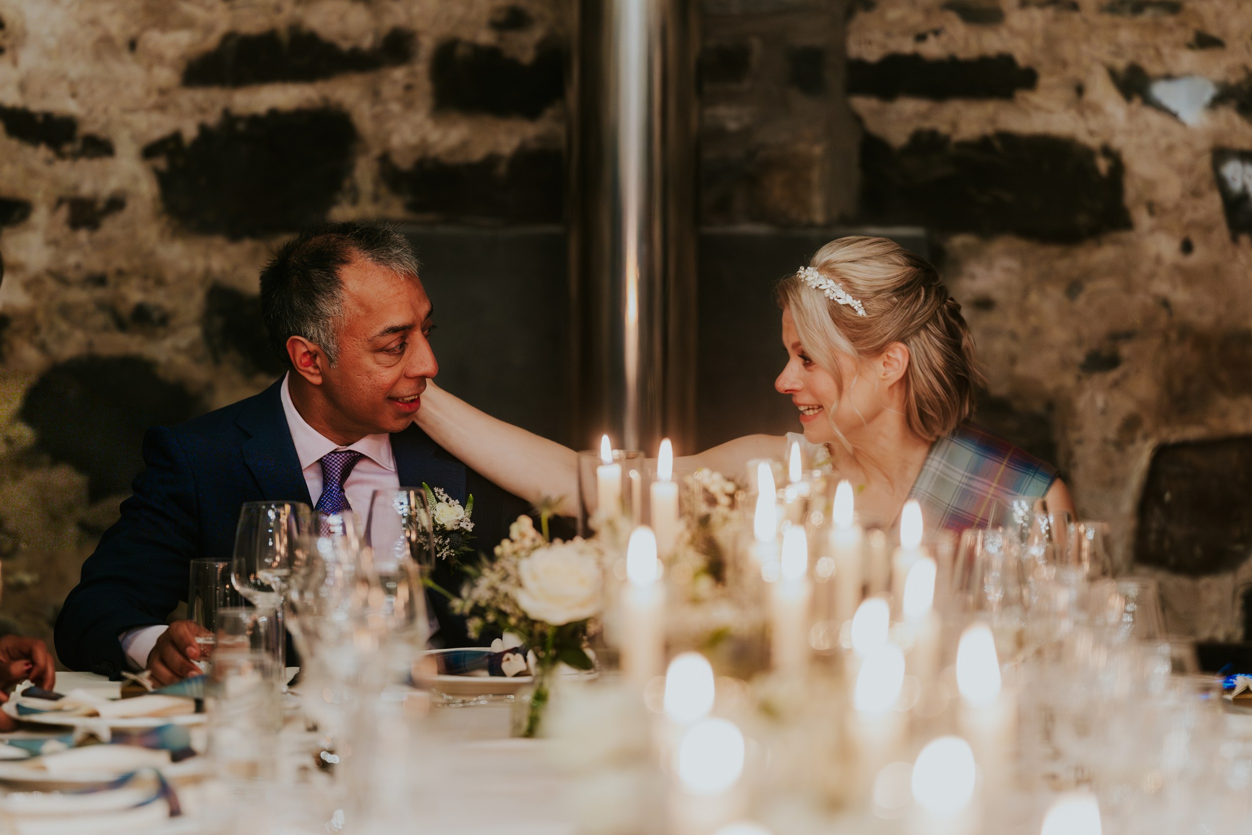 Lizanne and Jitin smile at each other across a candlelit head table in a stone-walled reception barn.