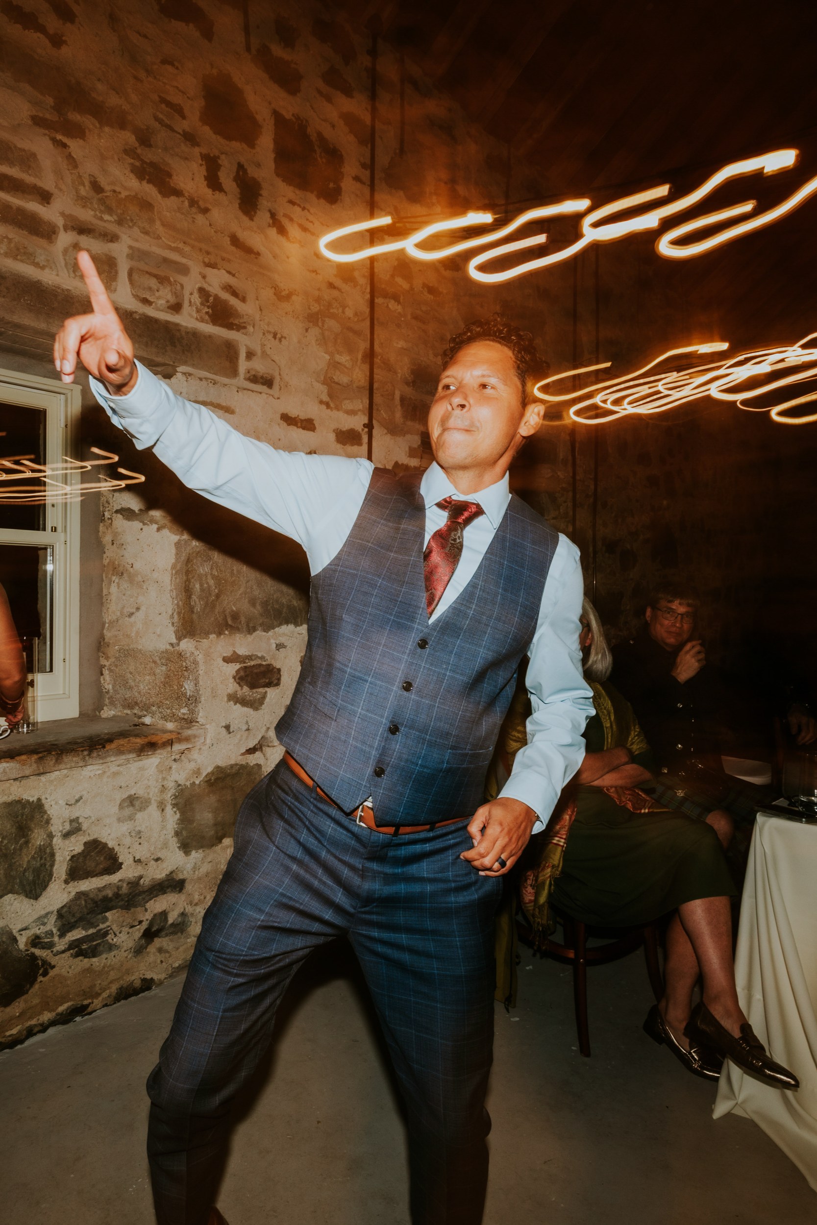 Male guest in a blue checked waistcoat dancing and pointing inside a stone-walled reception barn with streaked lights.