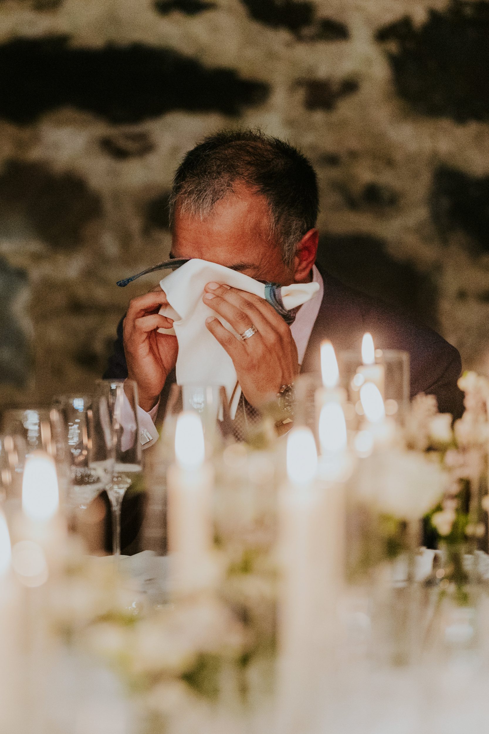 Man at reception table dabbing his eyes with a napkin among candles and glasses.