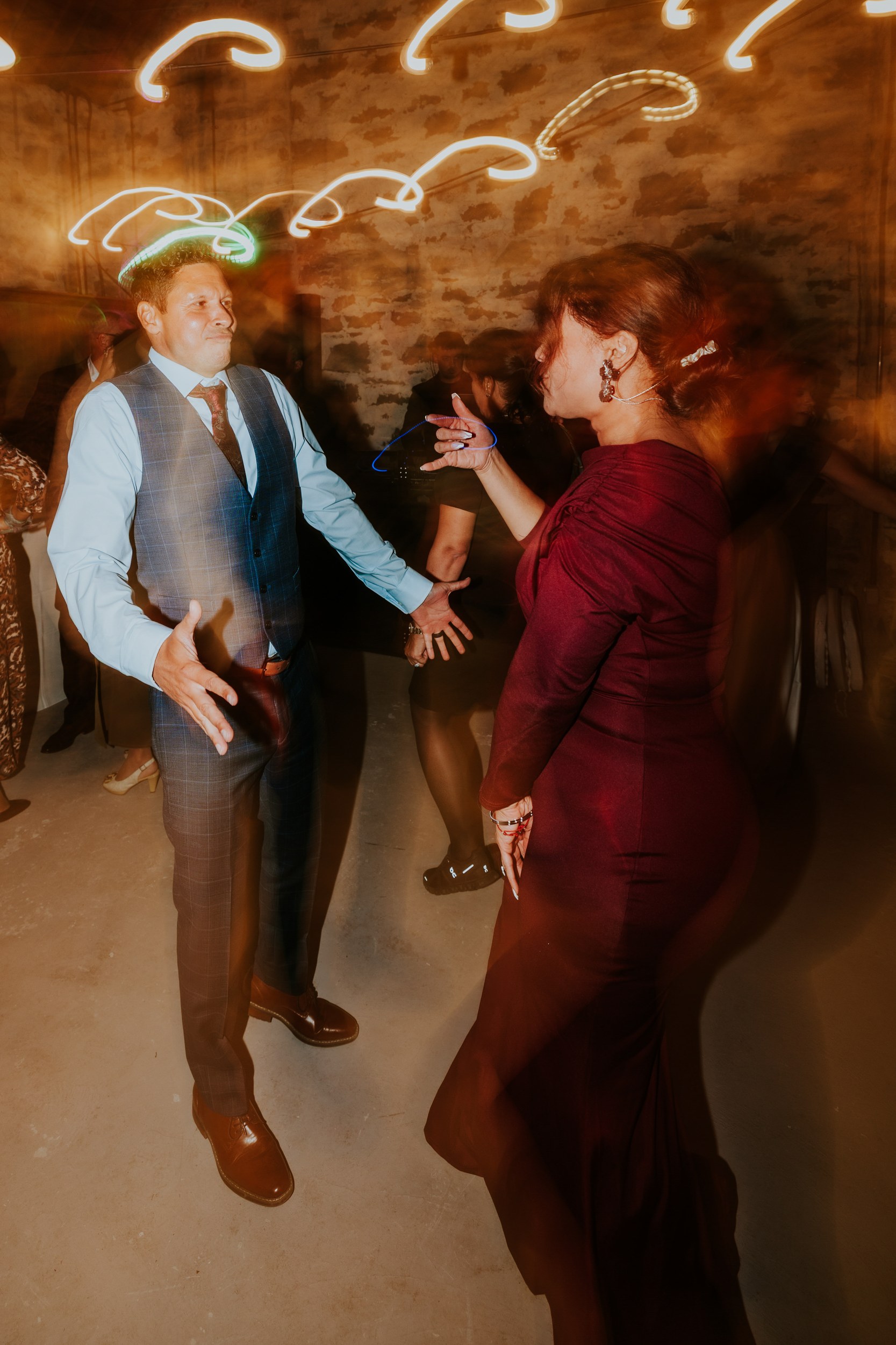 Man in waistcoat and woman in burgundy dress dancing in a stone-walled reception barn with light trail motion blur.