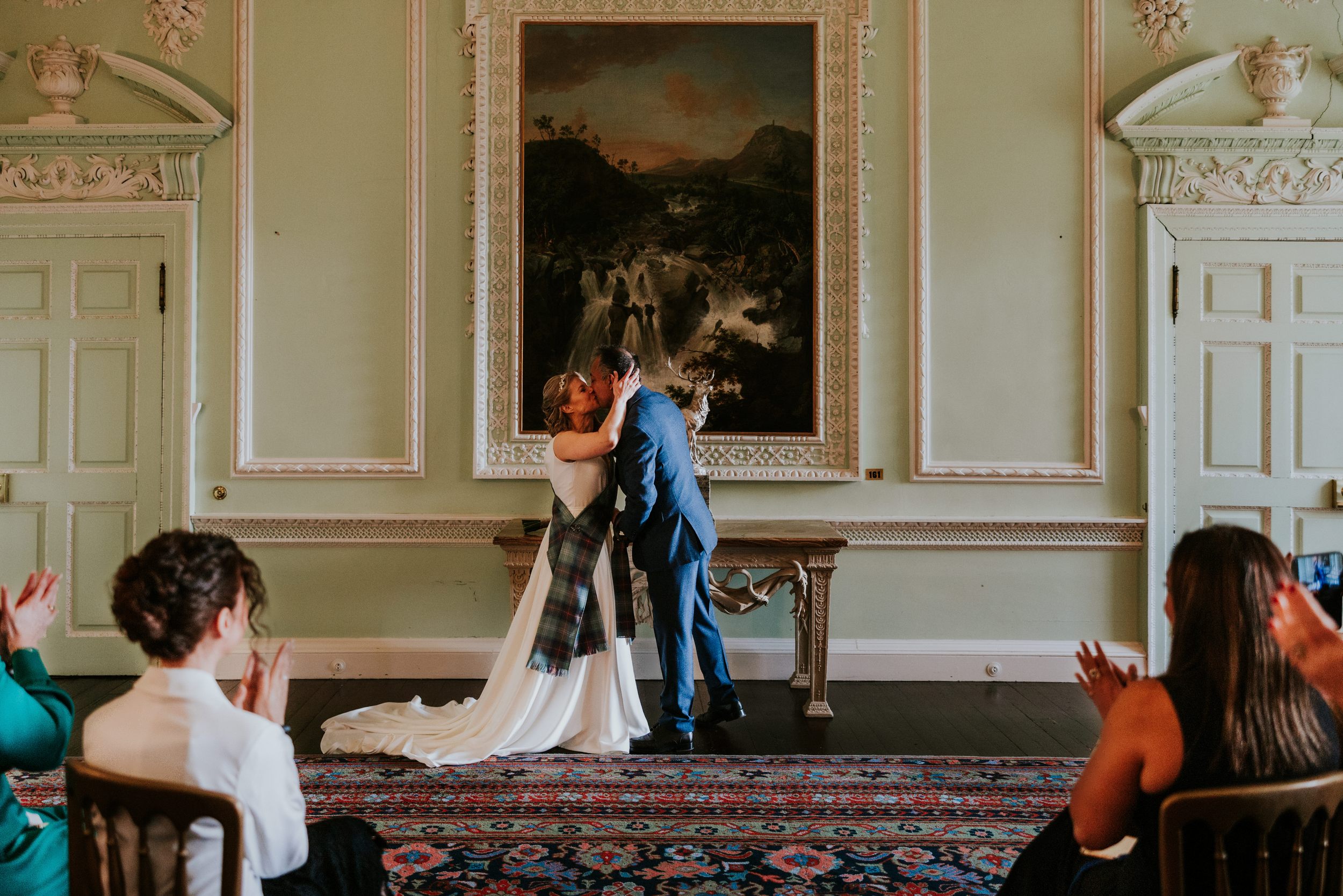 Newlyweds kiss after their ceremony in an ornate pale‑green room with a large painting, guests applauding.