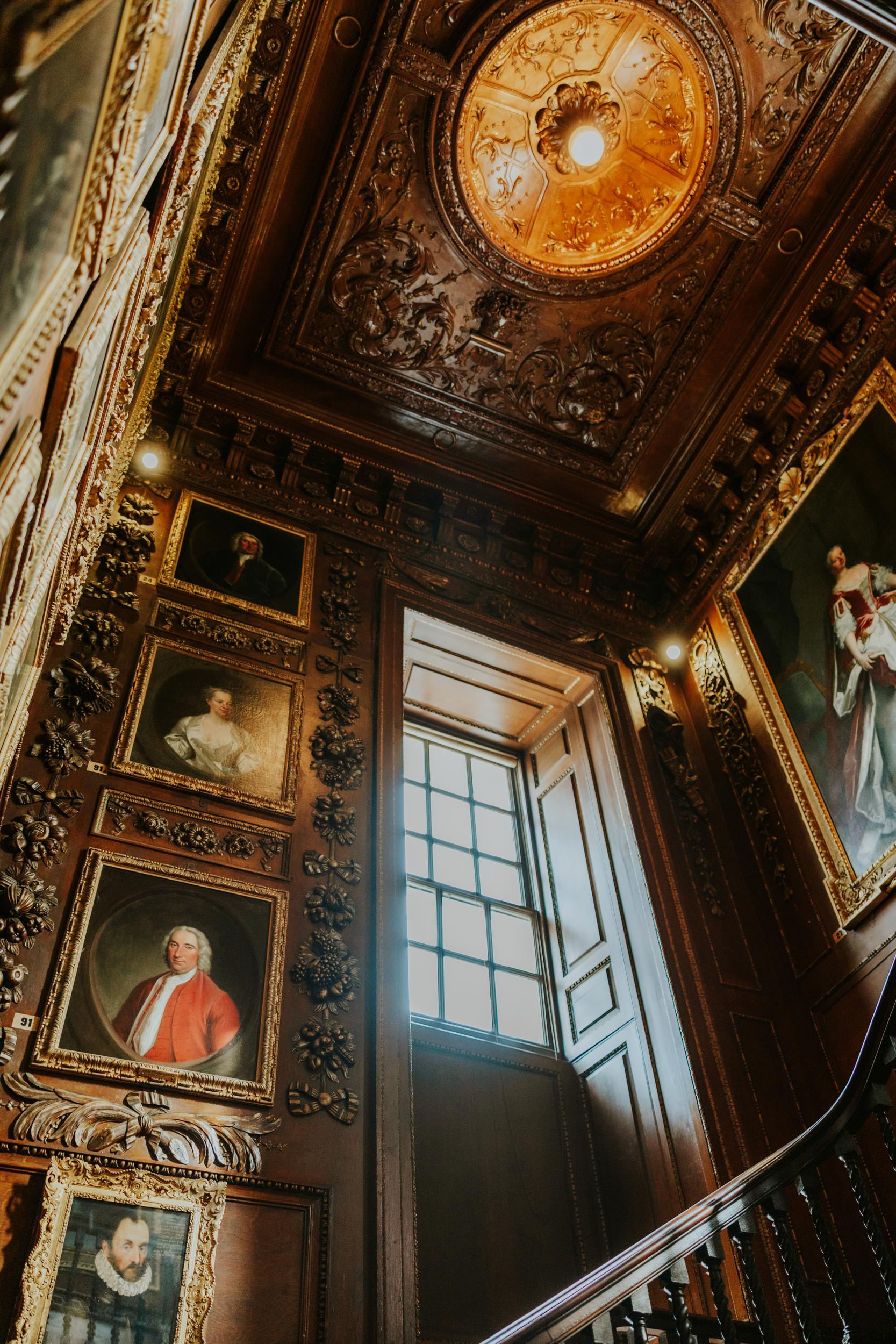 Ornate carved wooden ceiling, framed portraits, tall window and staircase inside Blair Castle.