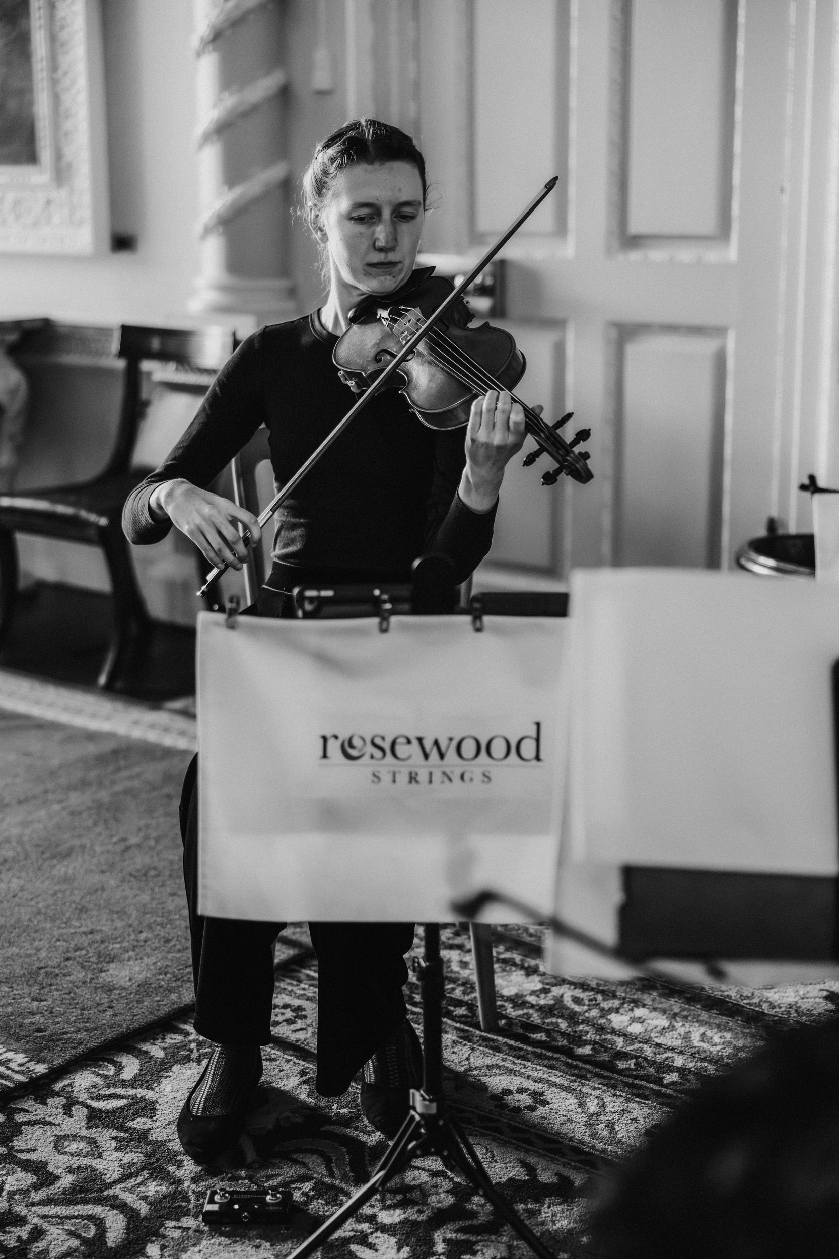 Seated violinist from Rosewood Strings playing indoors with music stand in a panelled room.