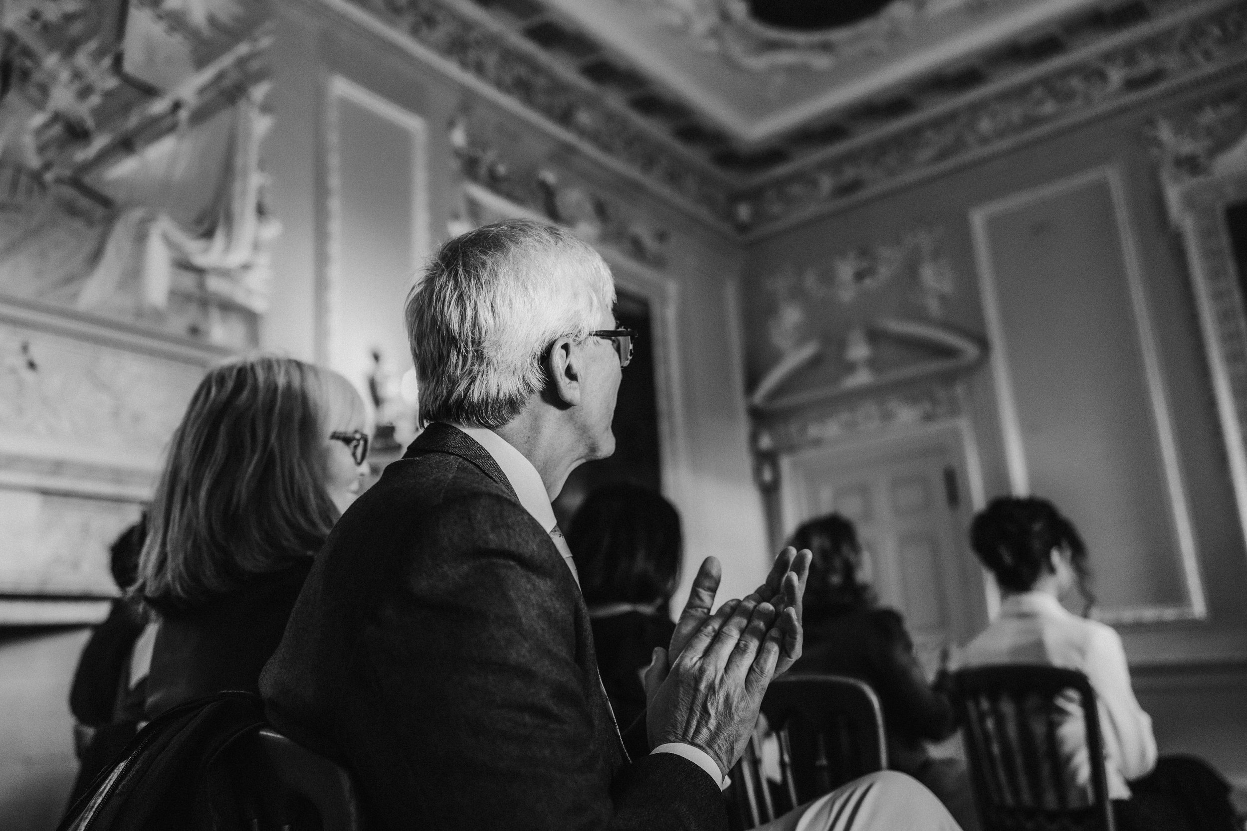Seated wedding guests in an ornate castle room, an older man with glasses clapping.