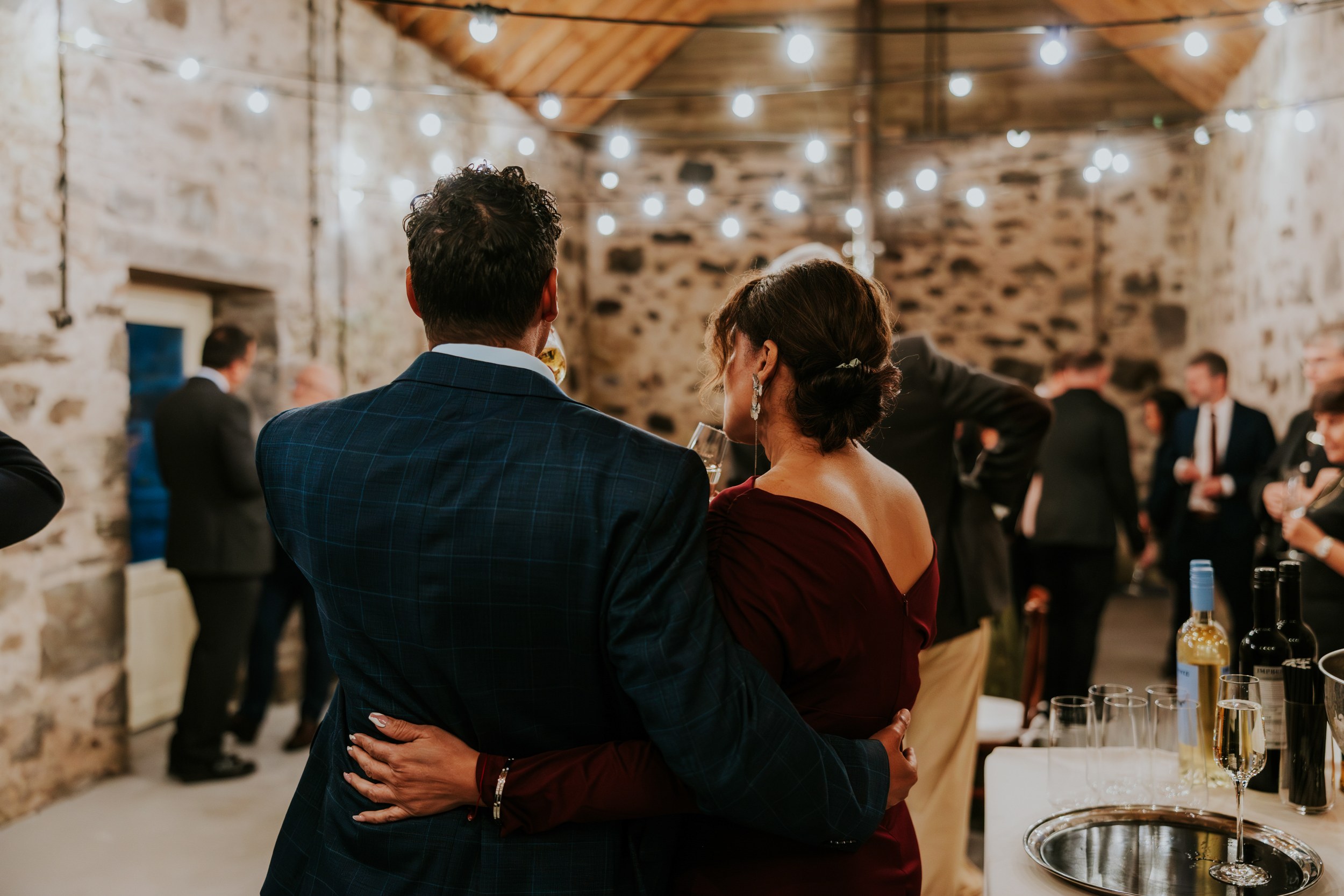 Two guests embracing at a stone-walled reception barn under string lights, with a drinks table to the right.